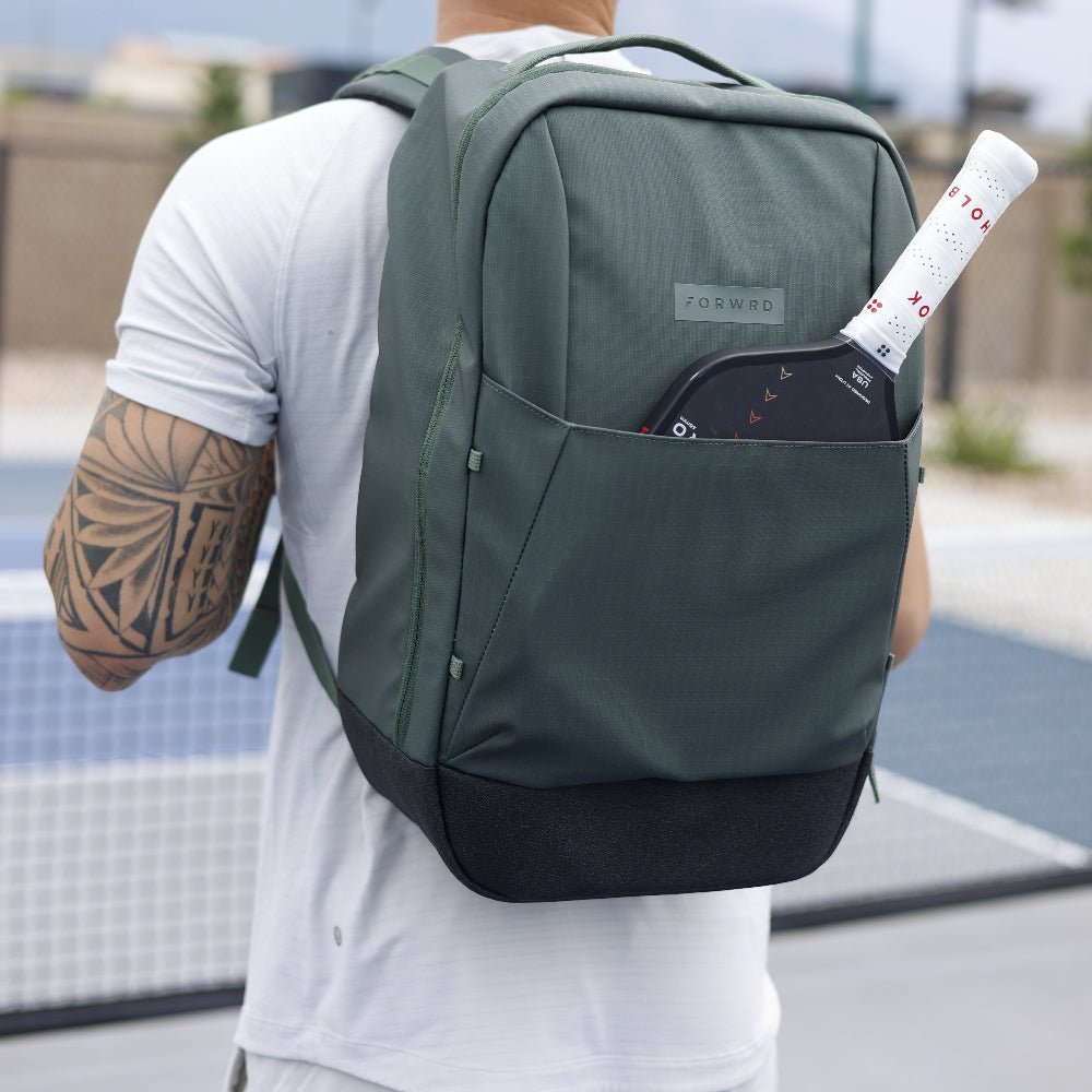 Dark green pickleball backpack by FORWRD, worn on a person's back, with a pickleball paddle in the side pocket and a black textured base.