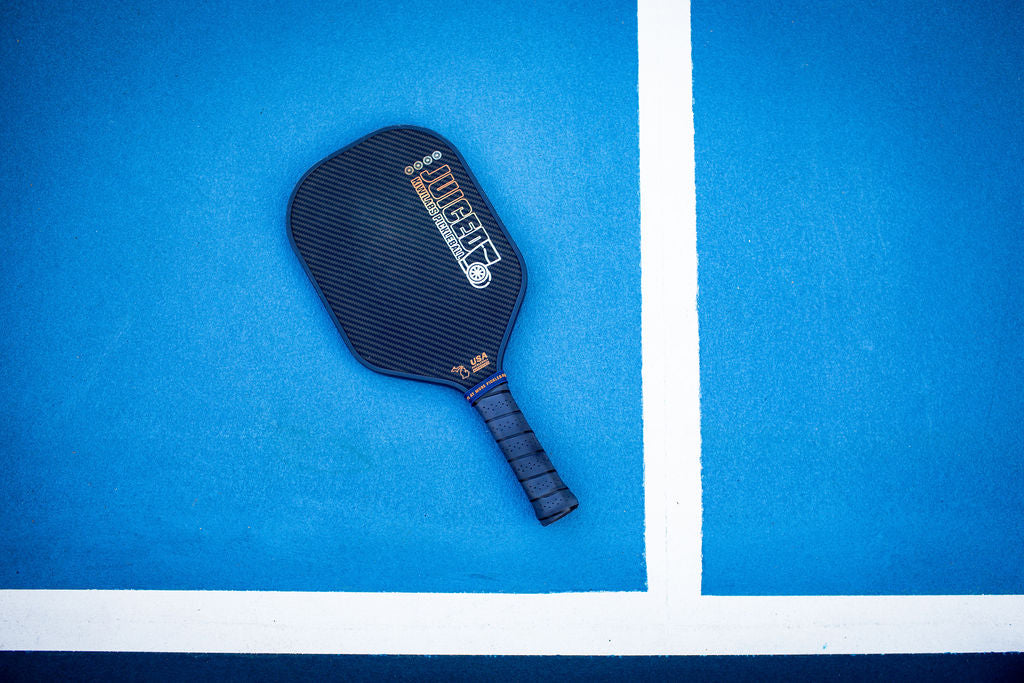 A black Juiced pickleball paddle resting on a blue court near a white line.