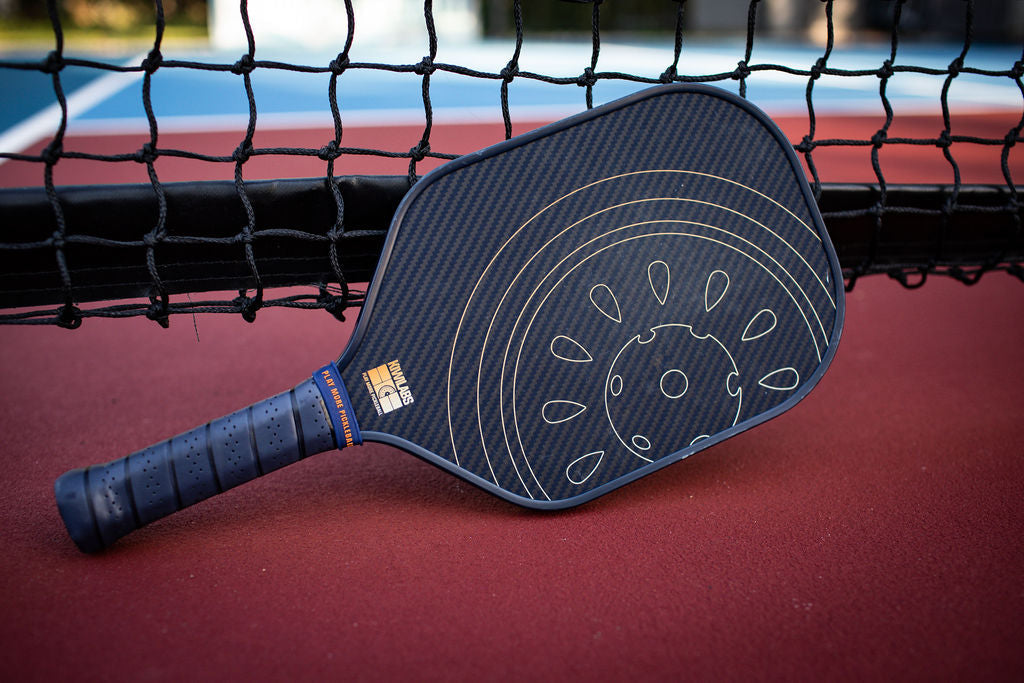 Close-up of a blue and black pickleball paddle resting on a red court against a black net.