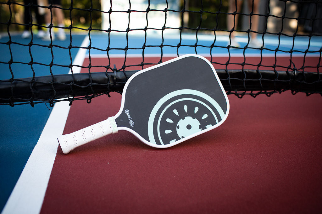 Close-up of a black and light blue pickleball paddle with a white grip resting on a red and blue court with a black net in the background.