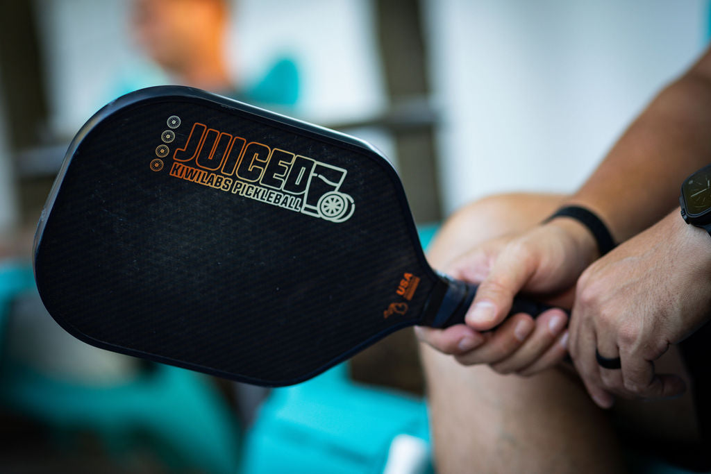 A person holding a black and orange Kiwi Labs Juiced pickleball paddle.