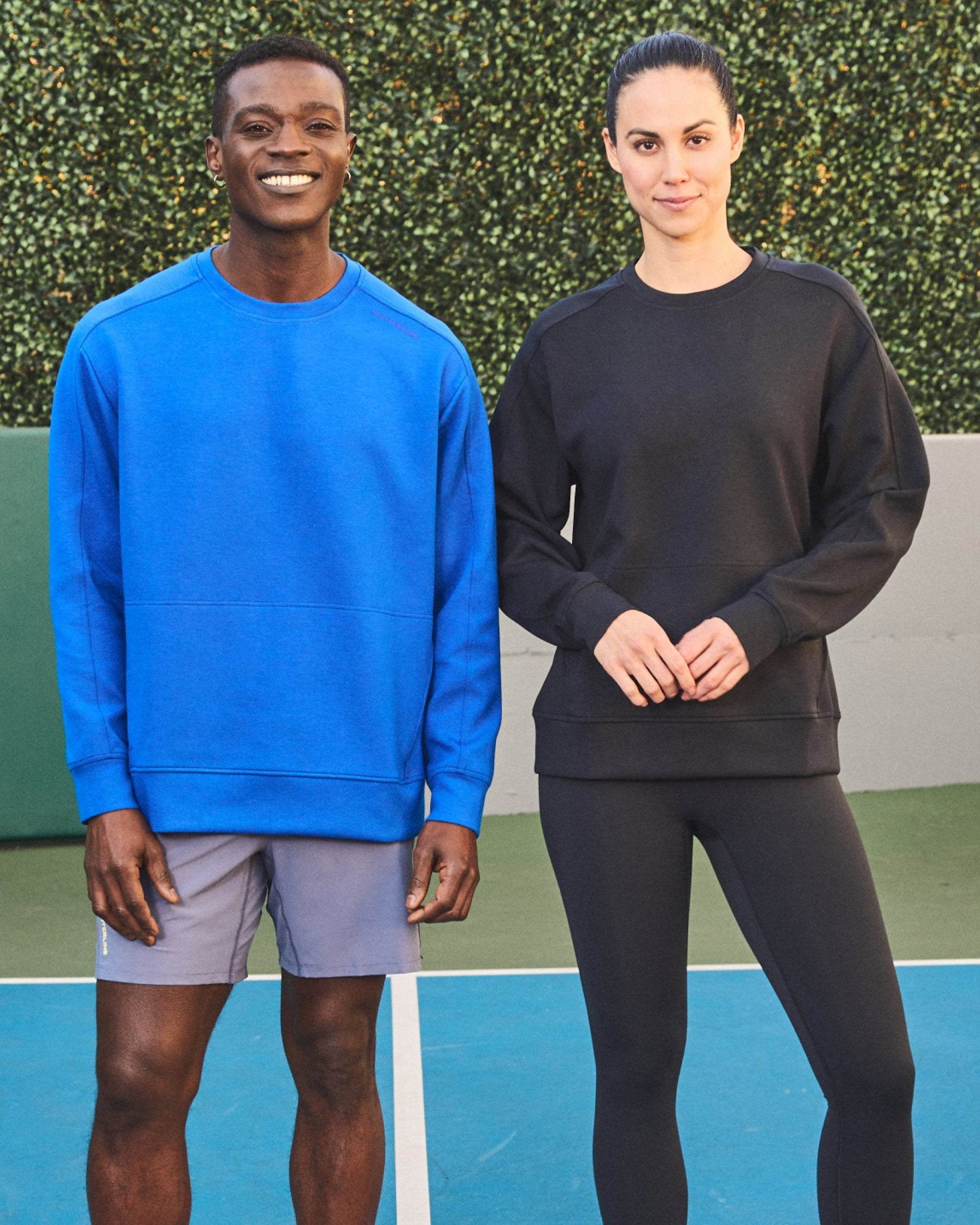 Two models in athletic wear stand against a green hedge. The man wears a blue long-sleeved shirt and gray shorts. The woman wears a black long-sleeved shirt and leggings.