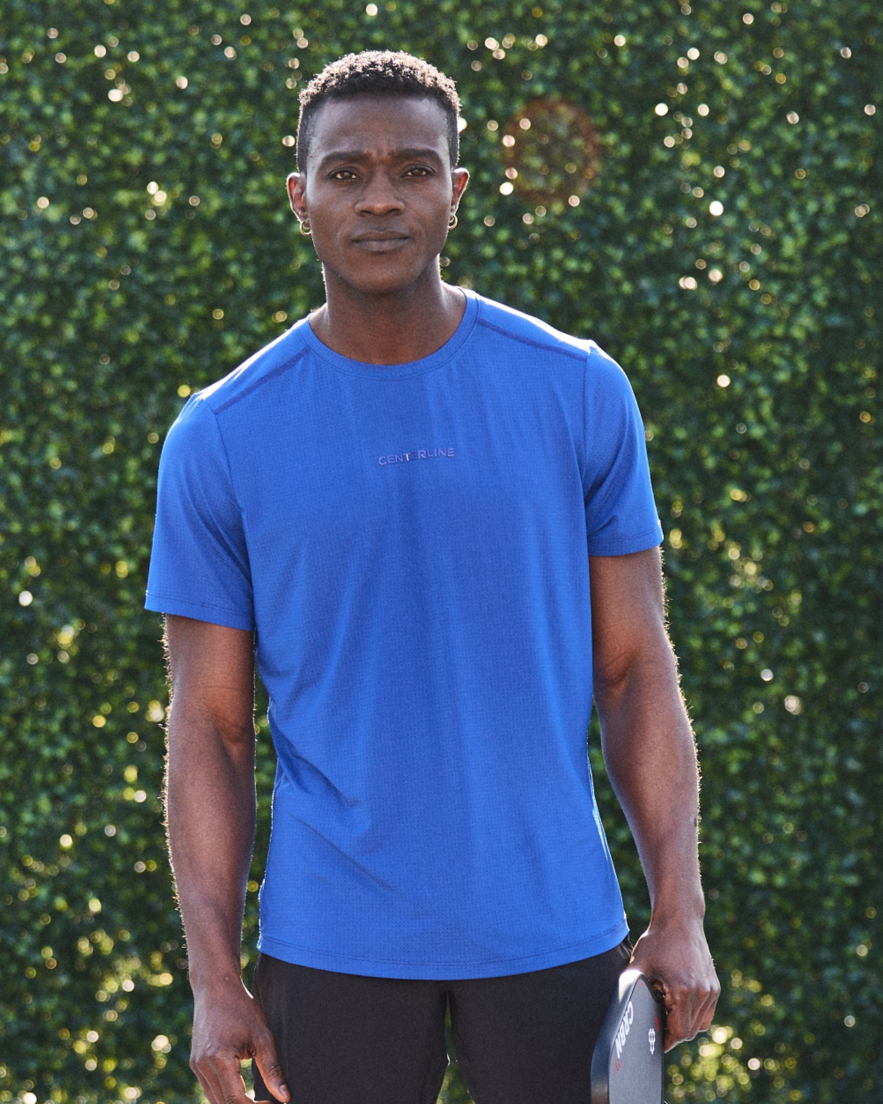 Man in blue athletic shirt with black shorts, holding a paddle in front of a green hedge.