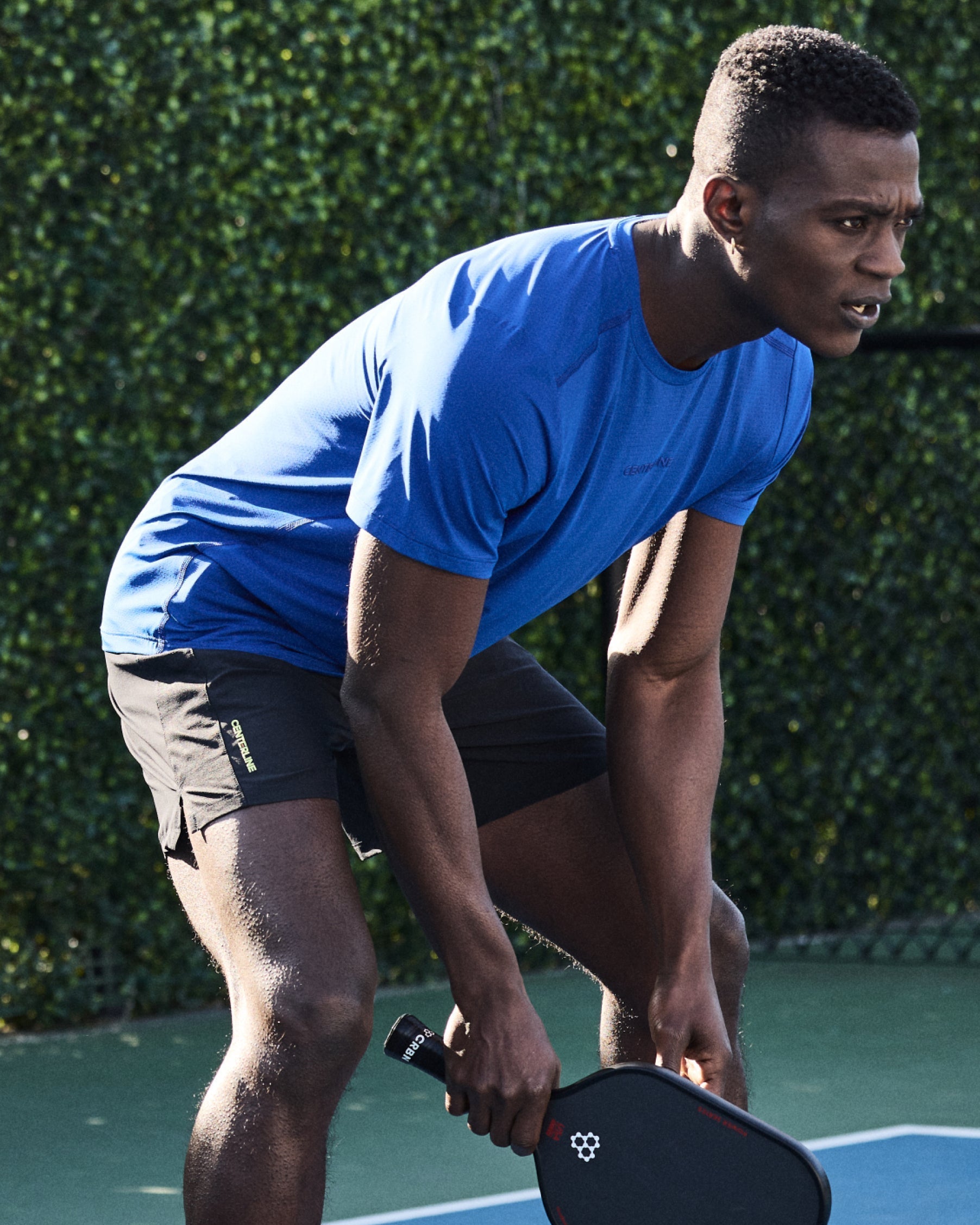 An athletic man in a blue shirt and black shorts leans forward, holding a pickleball paddle.