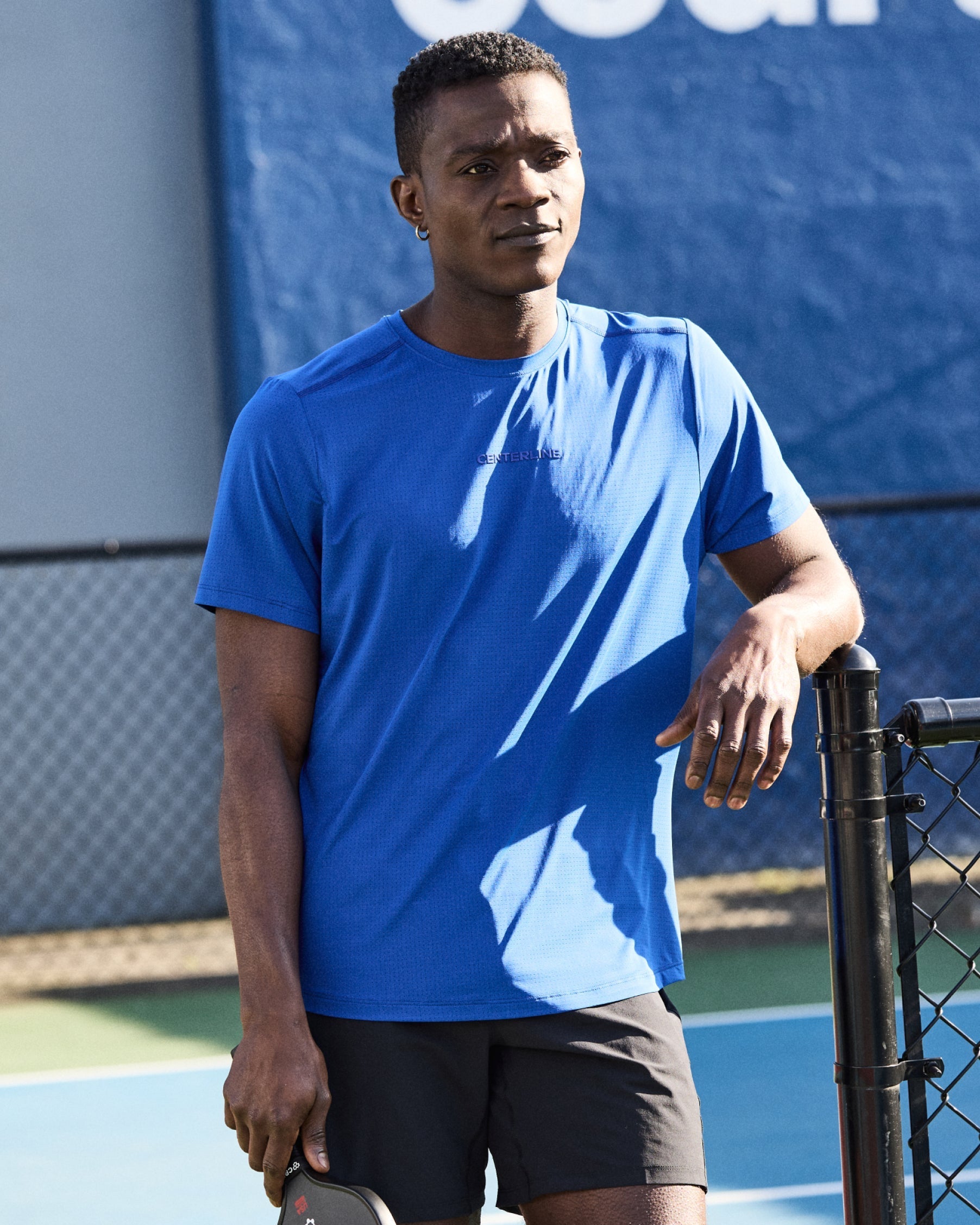 Man in blue shirt and black shorts leans on tennis court fence.