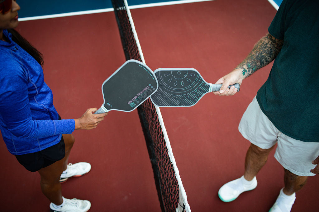 Overhead shot of two people holding pickleball paddles over a net on a maroon court, one person wears a blue top and black shorts while the other wears a green shirt and white shorts.
