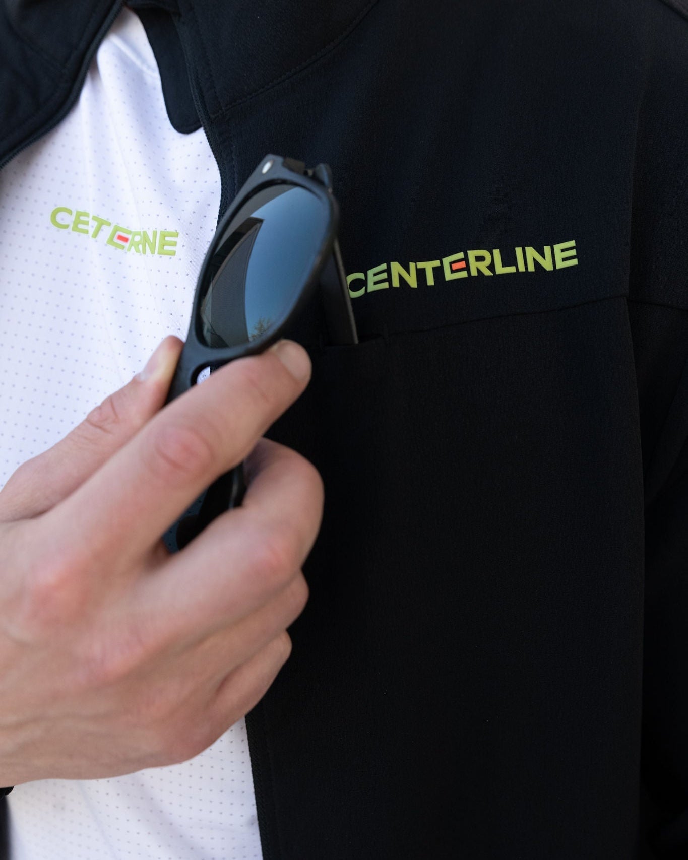 Close up of a person inserting sunglasses into the pocket of a black jacket with the word "CENTERLINE" in yellow and orange.