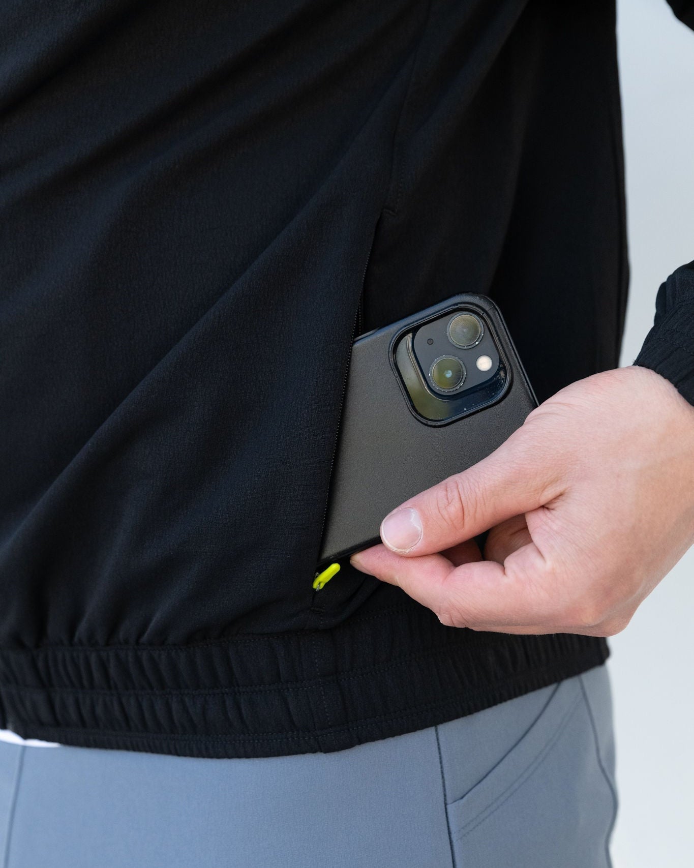 Person putting black smartphone in black jacket pocket. Gray pants.