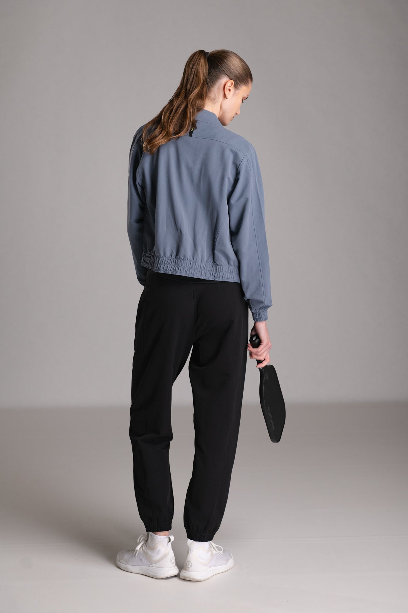 Woman from behind wearing a blue jacket, black pants, and white shoes, holding a pickleball paddle.