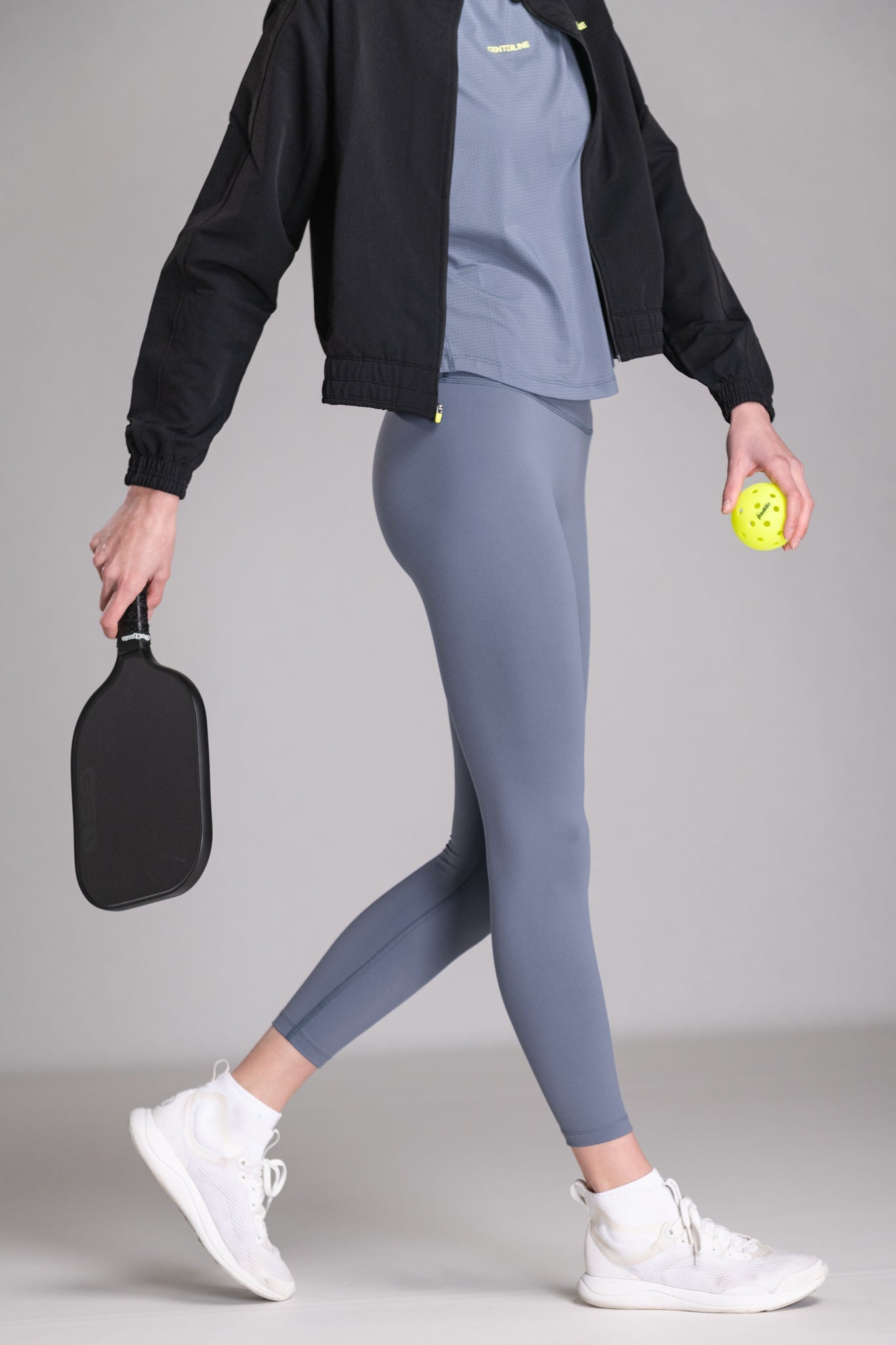 Woman wearing a black jacket, gray shirt and leggings, and white athletic shoes, holding a pickleball paddle and ball.