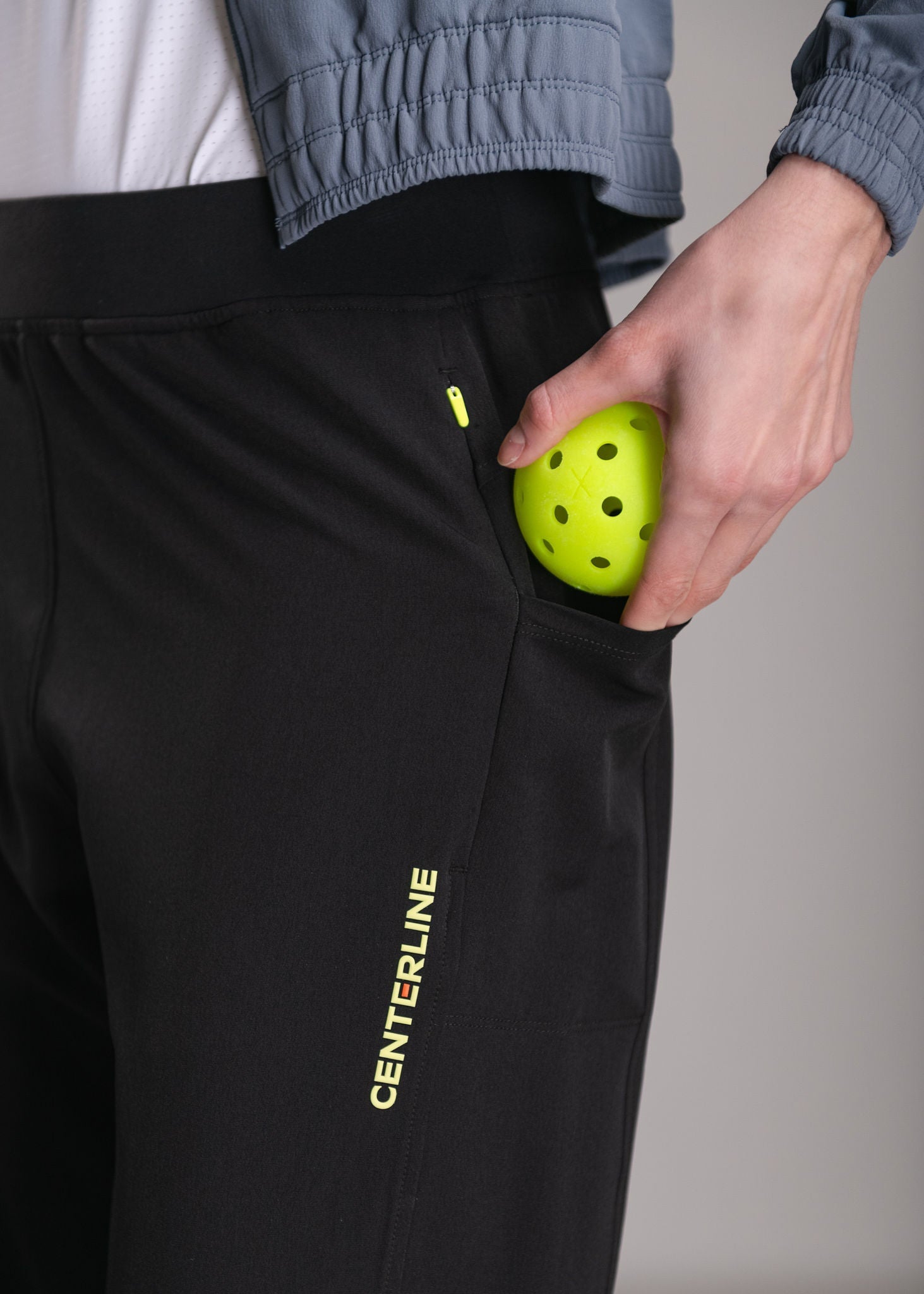 Close-up of black athletic pants with a neon yellow pickleball in the pocket, and a matching jacket. The word "CENTERLINE" is printed vertically on the pants' leg.