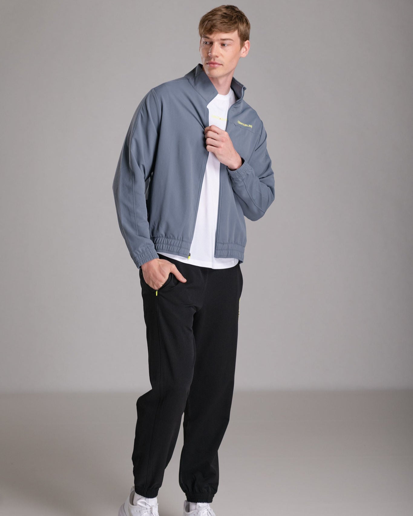 Man wearing a blue-grey jacket over a white shirt, paired with black sweatpants and white sneakers.