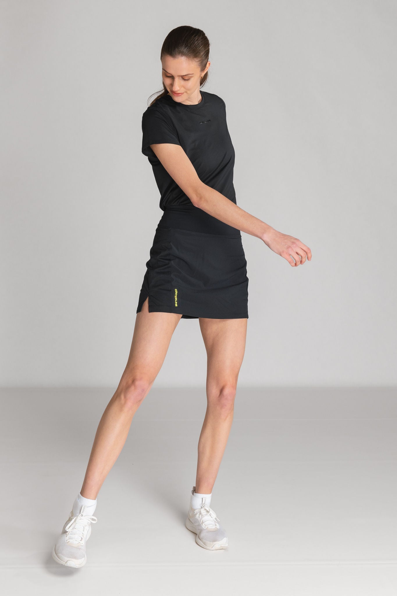 A woman in a black T-shirt and a black athletic skirt with yellow lettering poses in white sneakers.