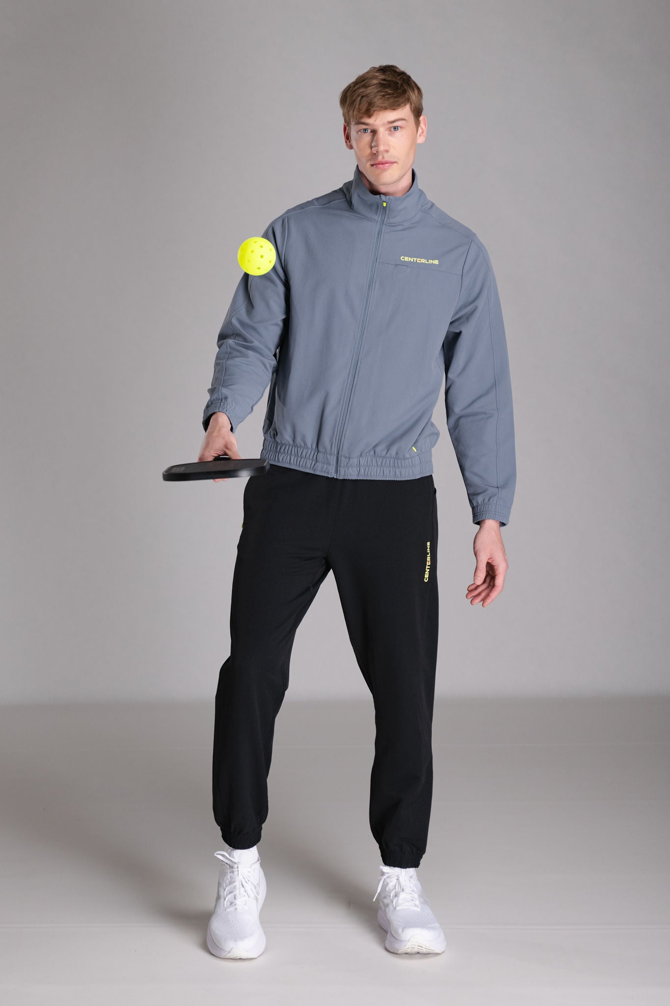 Man in gray jacket and black pants holding a pickleball paddle and ball.