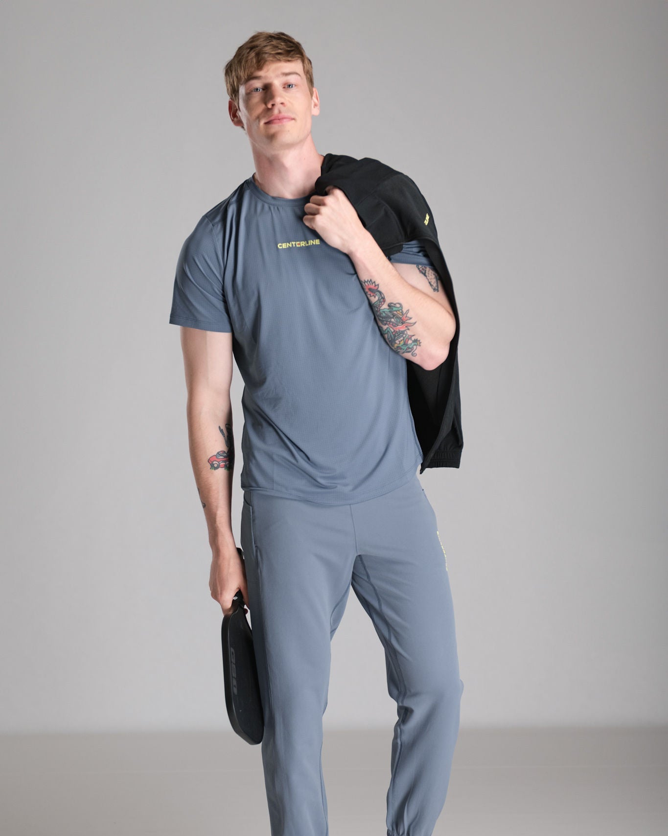 Man wearing gray activewear set with tattoos holding a paddle and jacket against a gray backdrop.