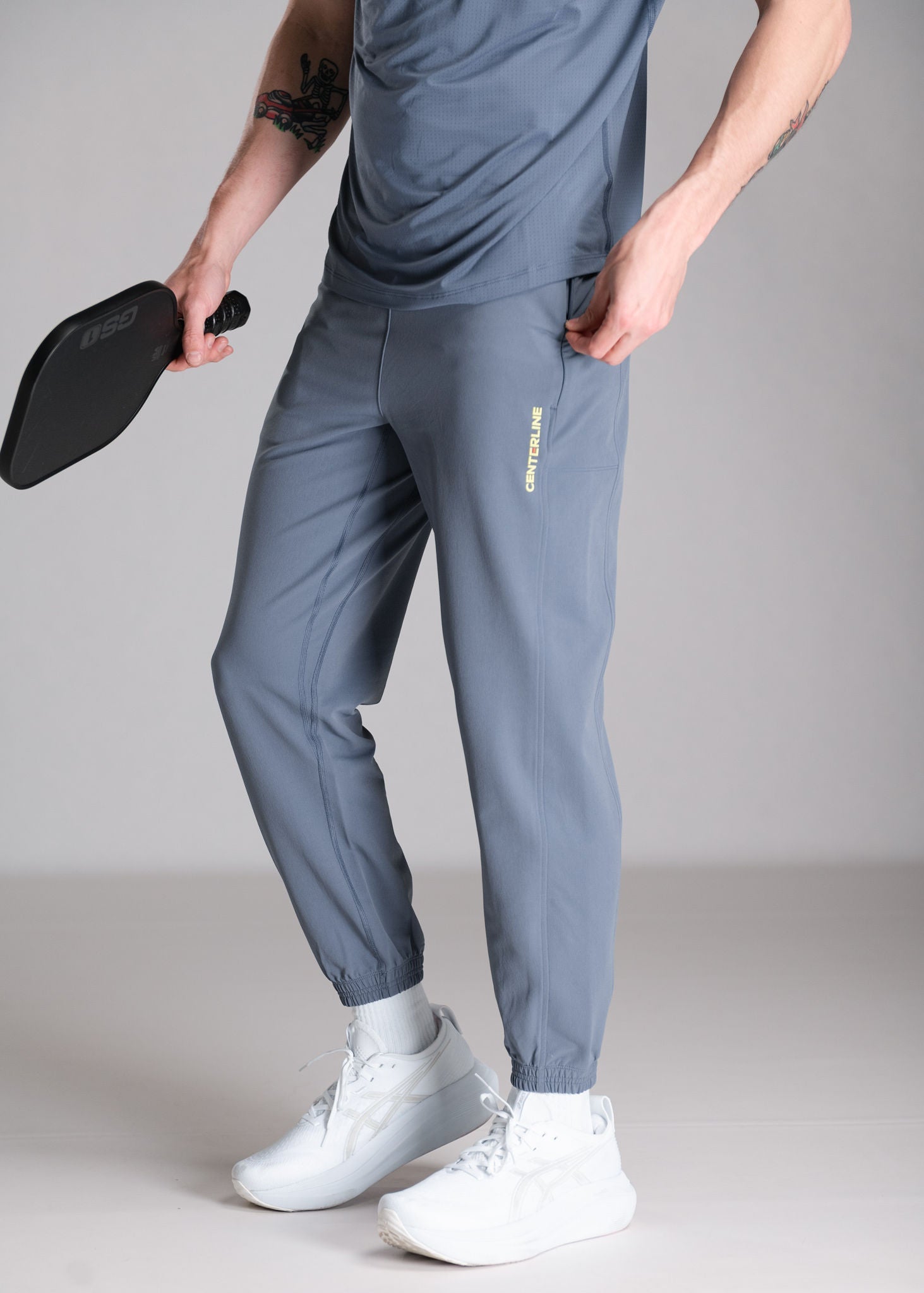 A person wearing gray athletic pants and a matching shirt, holding a black paddle and white athletic shoes, posing against a gray backdrop.