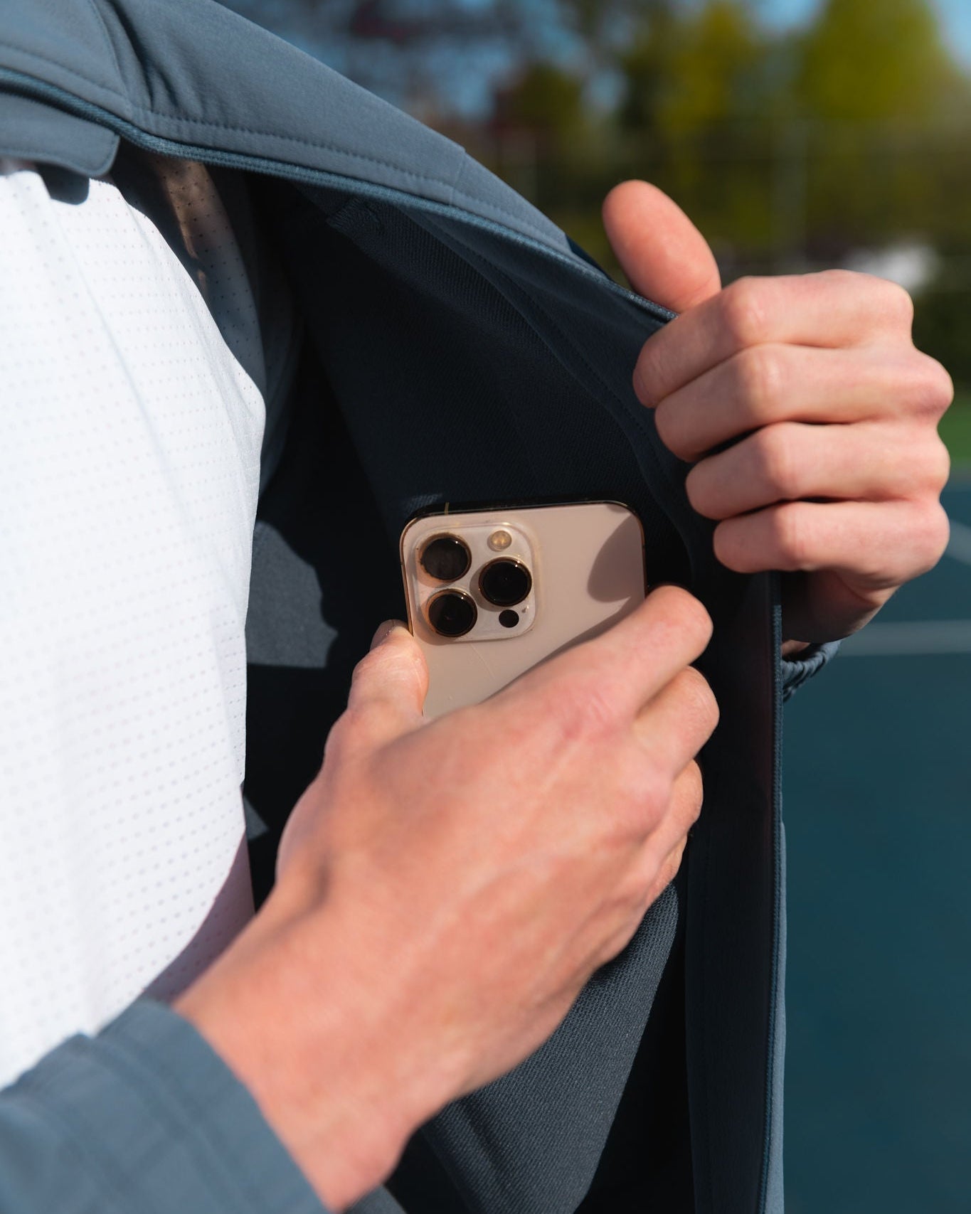Person placing a gold iPhone into a blue-grey jacket pocket, wearing a white shirt.