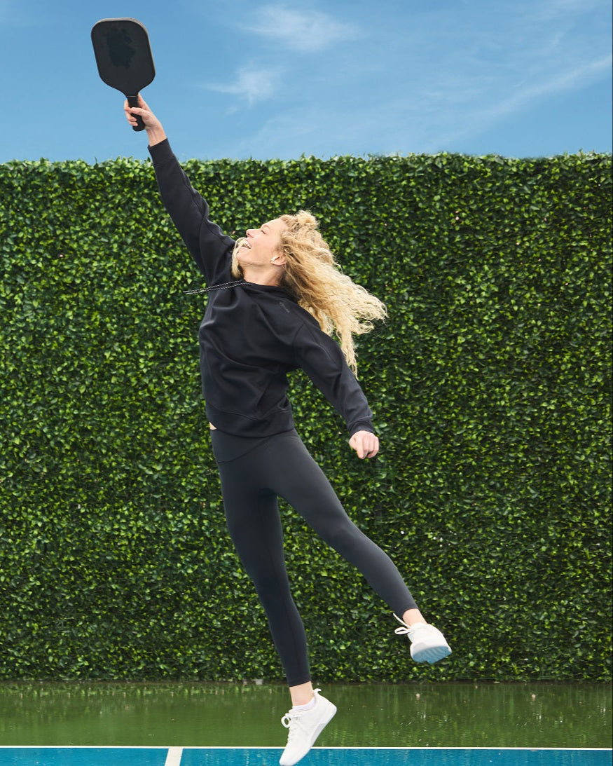 Woman in black athletic wear jumping with pickleball paddle in hand.