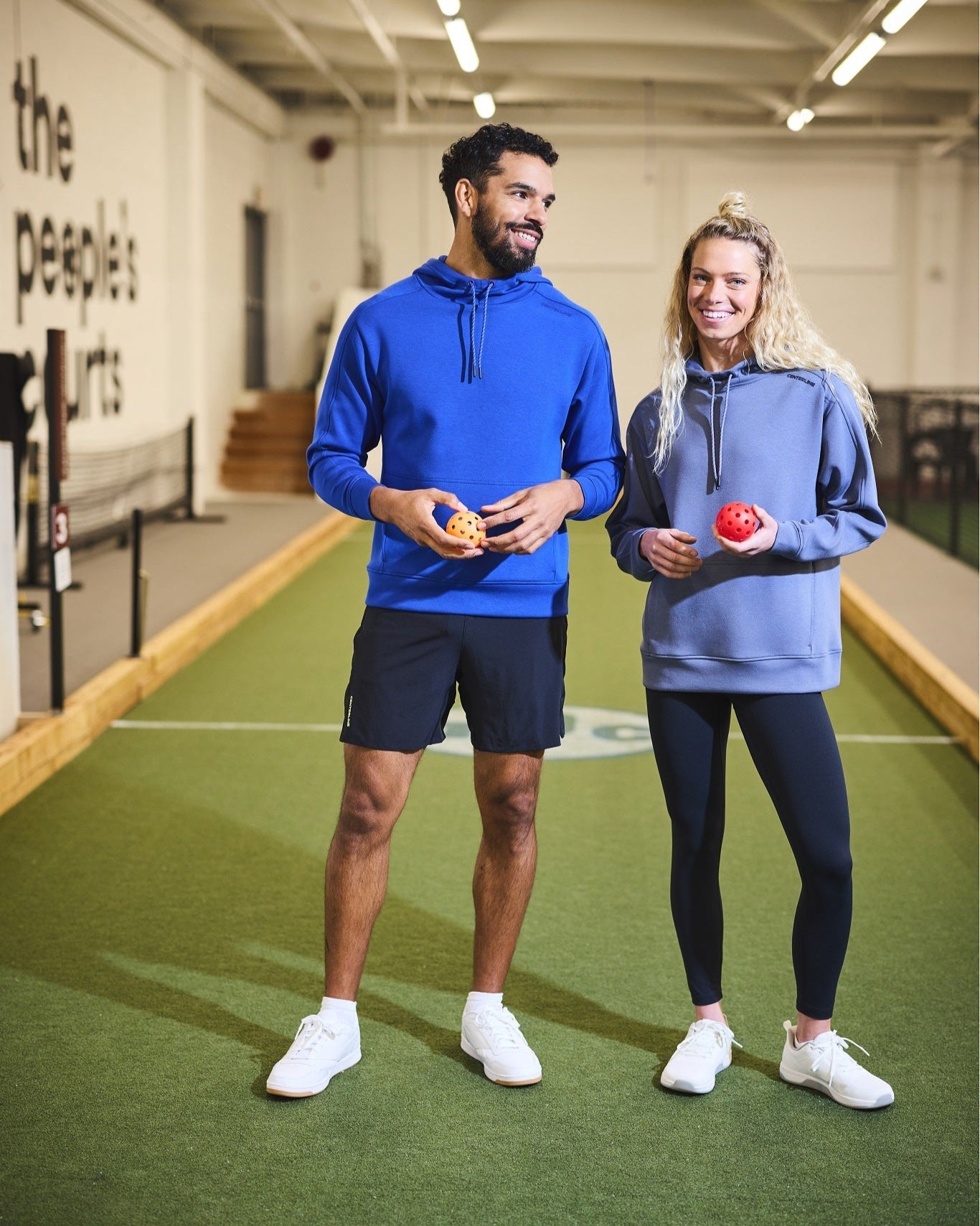 Two people standing on a bocce court in matching hoodies and athletic shoes.
