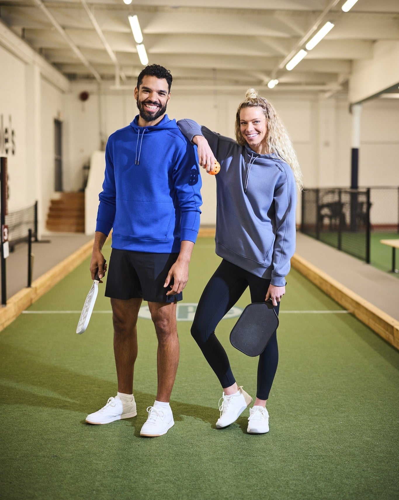 Two people wearing hoodies holding pickleball paddles standing on a pickleball court.