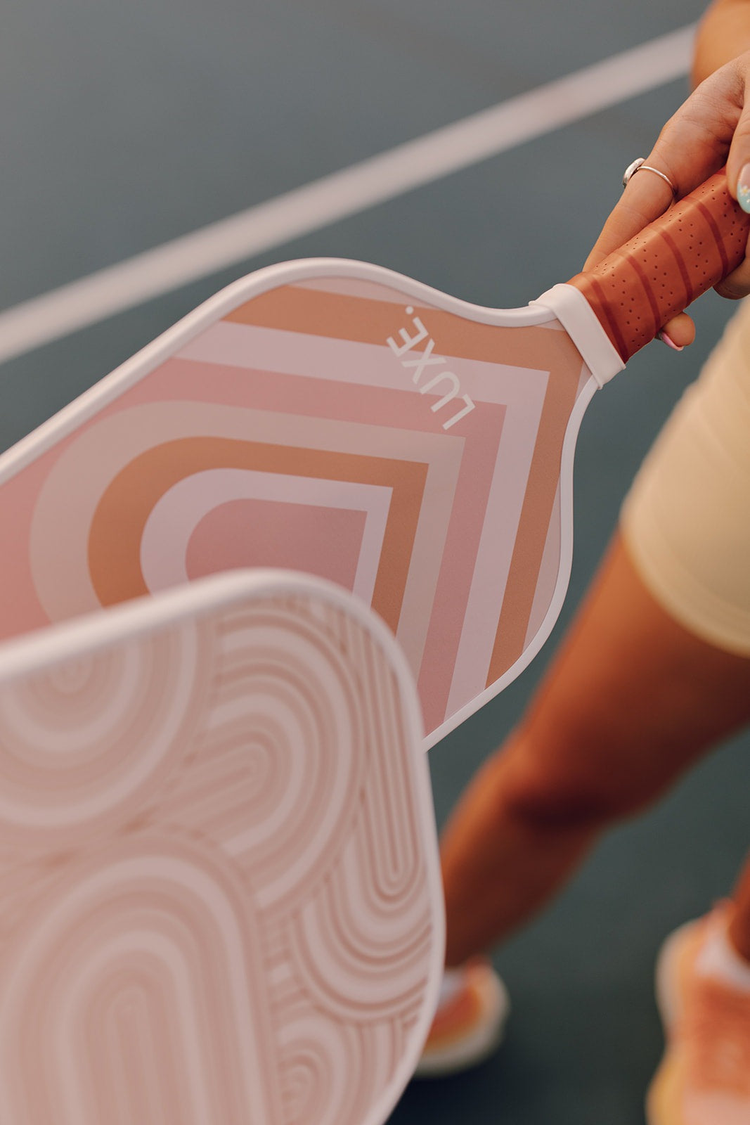 A woman holds two pink pickleball paddles, one with horizontal stripes and the word "LUXE." The paddles have white edges and tan handles. She is wearing a yellow athletic outfit and orange sneakers. The paddles are on a pickleball court.