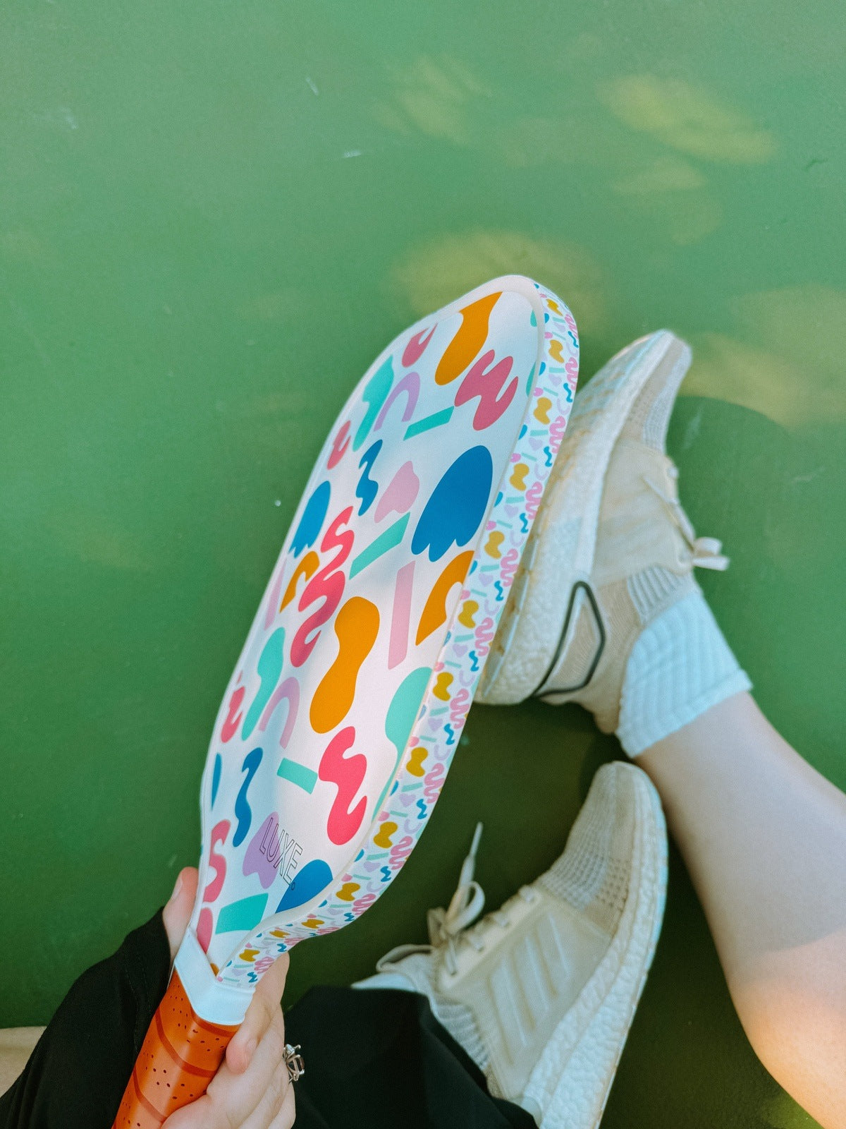 Overhead shot of a person holding a pickleball paddle with colorful abstract shapes while wearing light-colored sneakers on a green surface.
