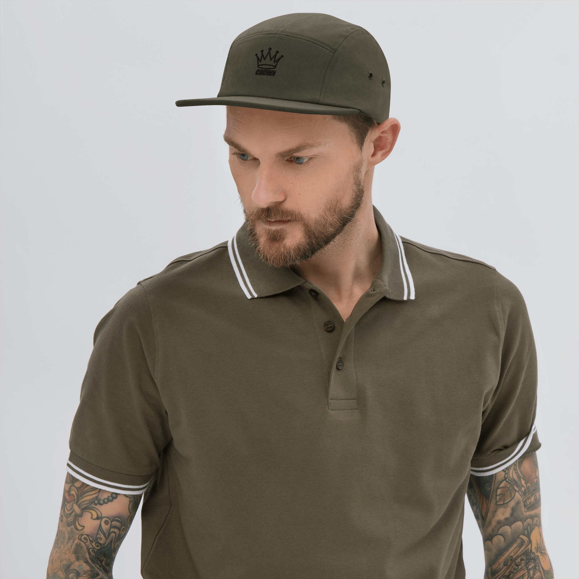 Man wearing olive green polo shirt and matching cap, looking down. Tattoos on arms.