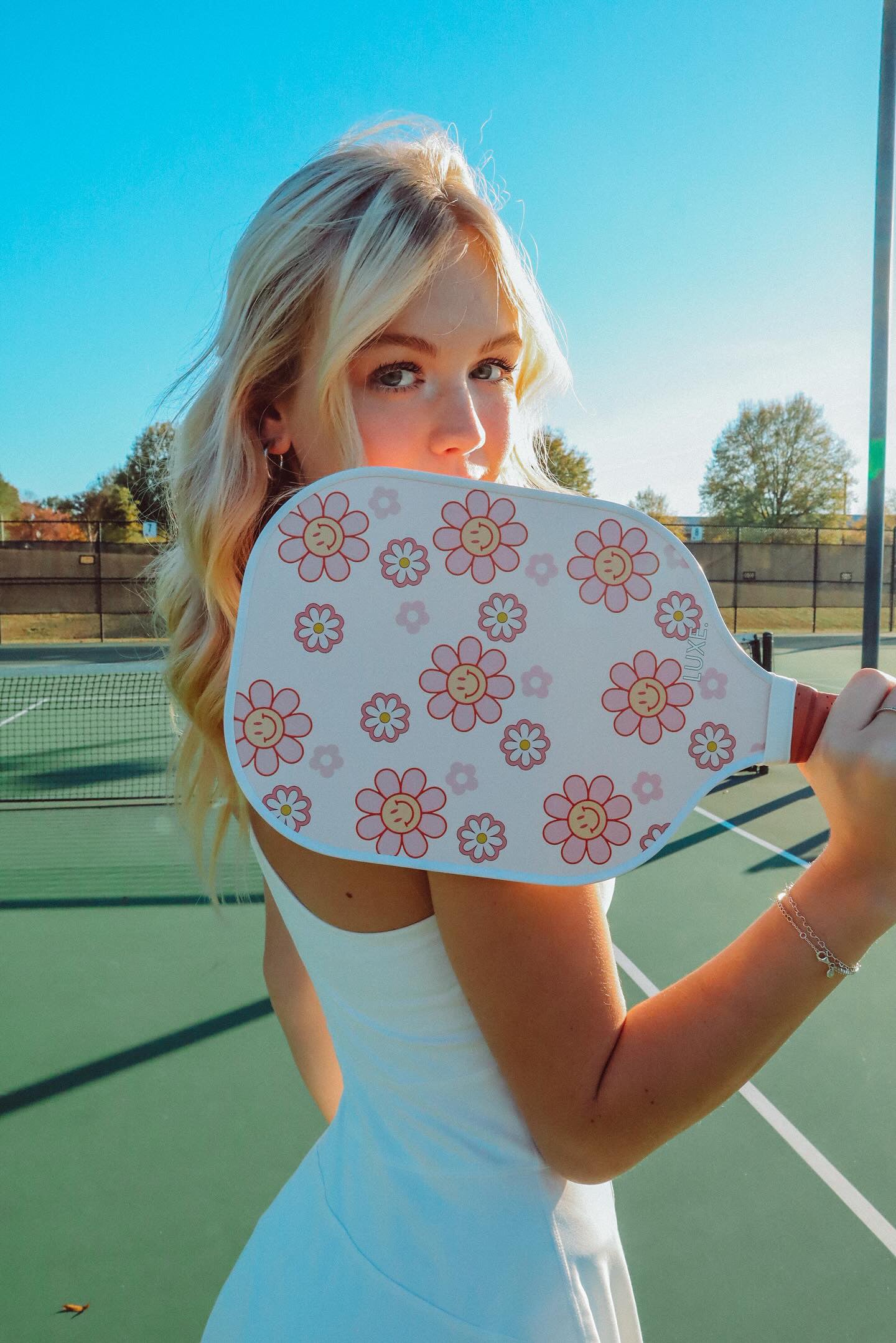 Blonde woman in white dress holding floral pickleball paddle on green tennis court.