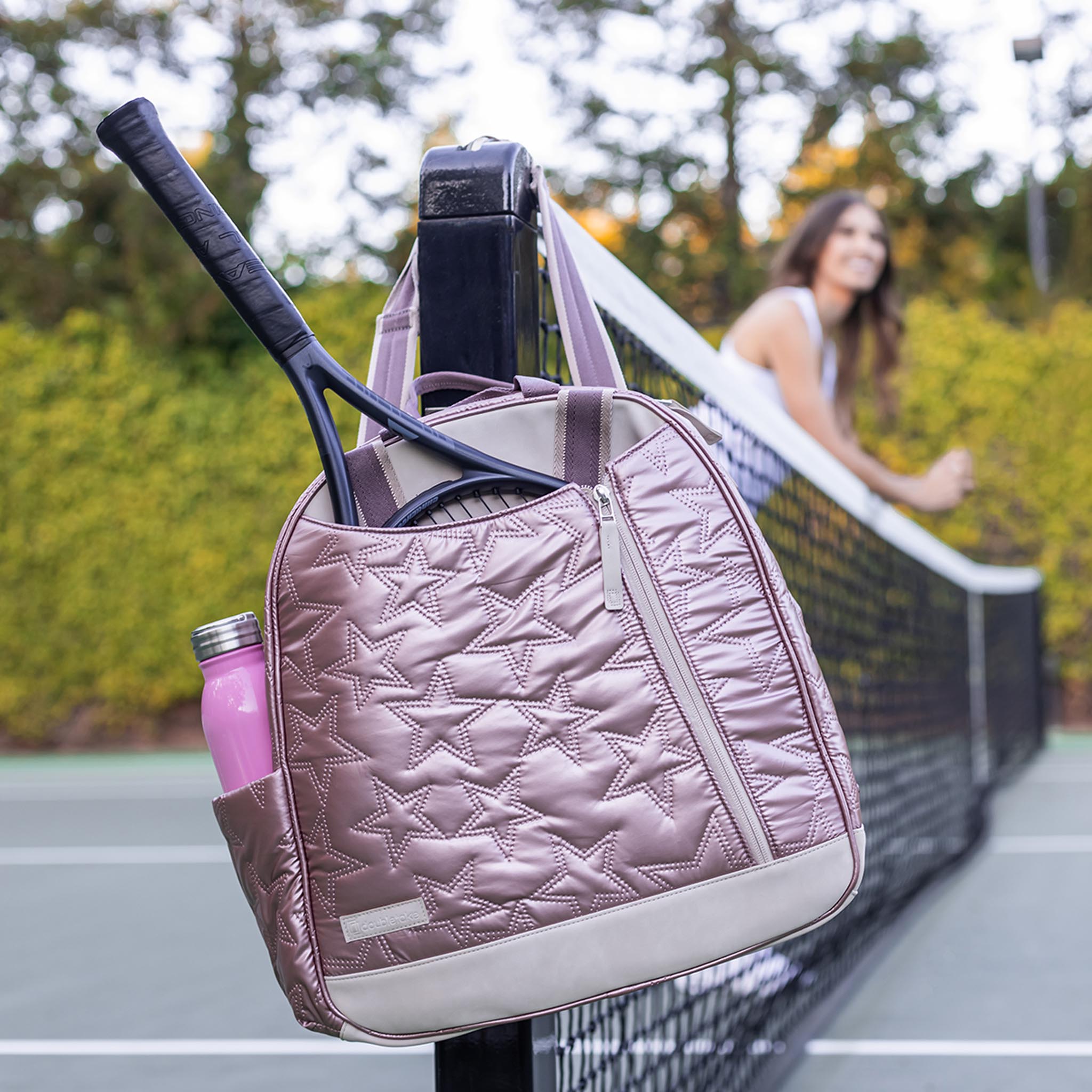 Rose gold quilted tennis backpack by Doubletake with star pattern, holding a racket and pink water bottle, hanging on a court net.
