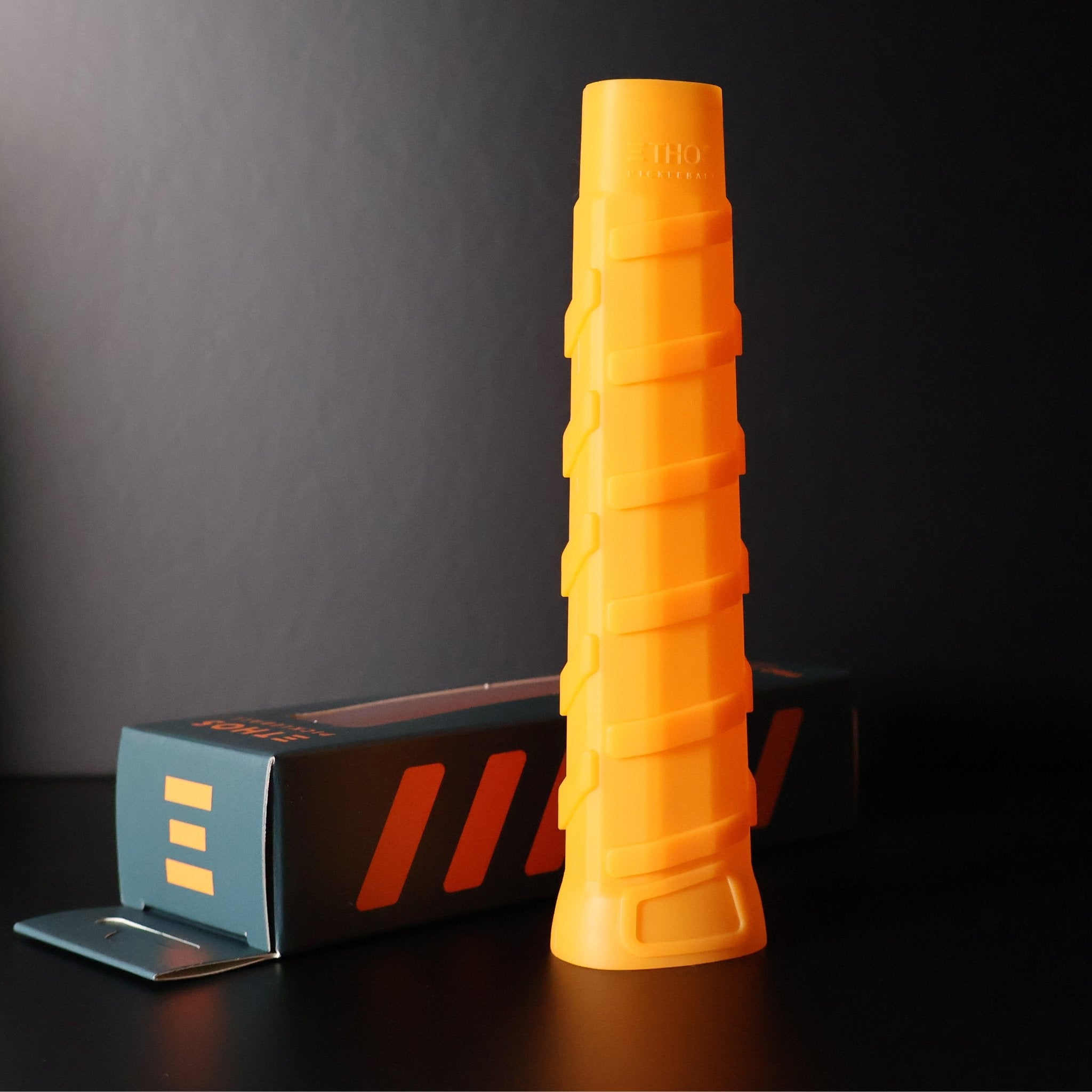 Vibrant orange ribbed Pro Undergrip by Ethos Pickleball, with logo at top, standing beside its green and orange packaging box.