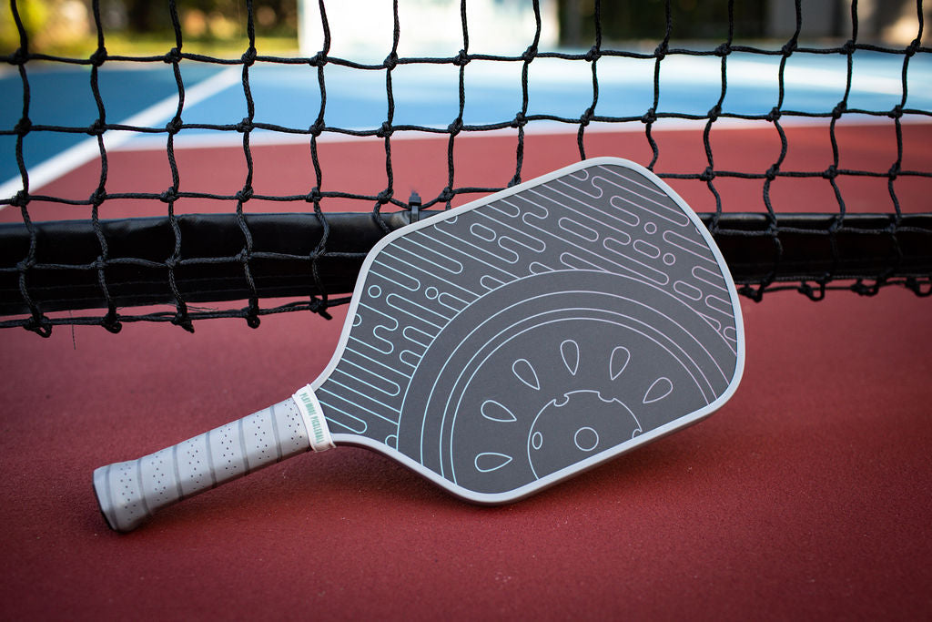 Close-up of a gray and white pickleball paddle with a decorative design, resting on a red pickleball court against a black net.
