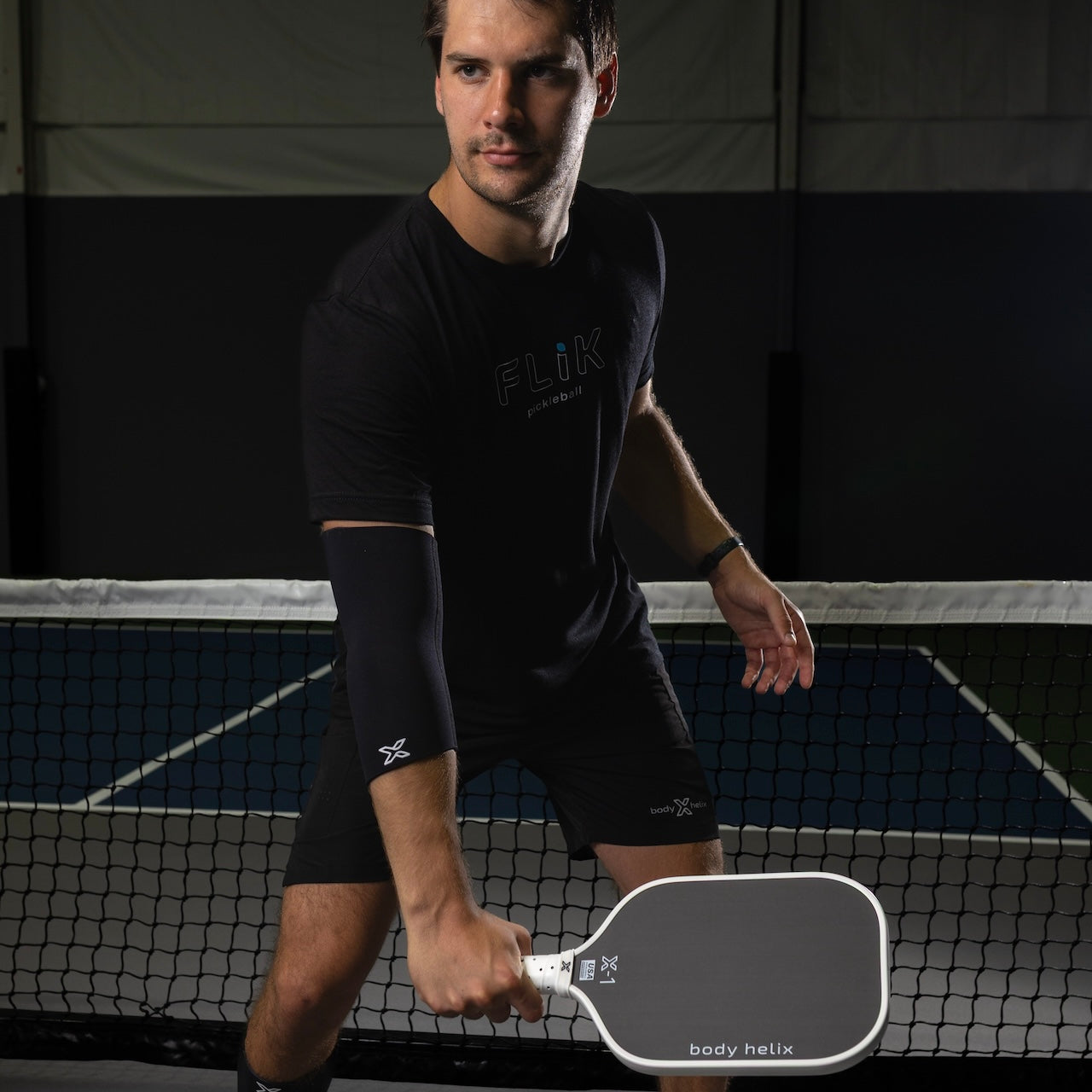 Man in black t-shirt and shorts holding a pickleball paddle on an indoor court.