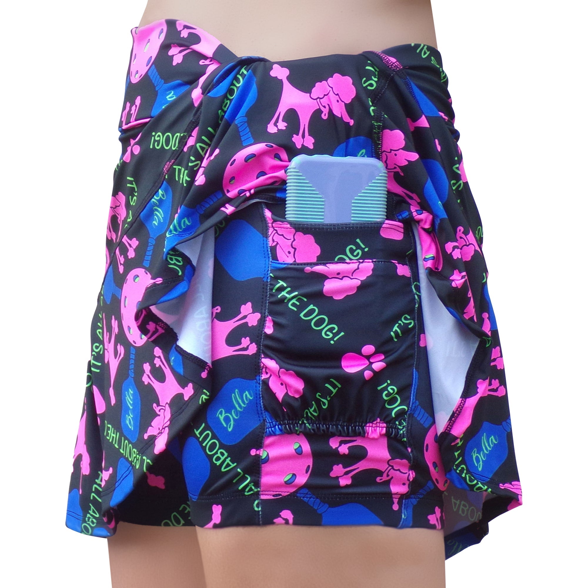 Black A-line skort by Pickleball Bella, with pink poodle and pickleball patterns, 'IT'S ALL ABOUT THE DOG!' text, and a blue case in its side pocket.
