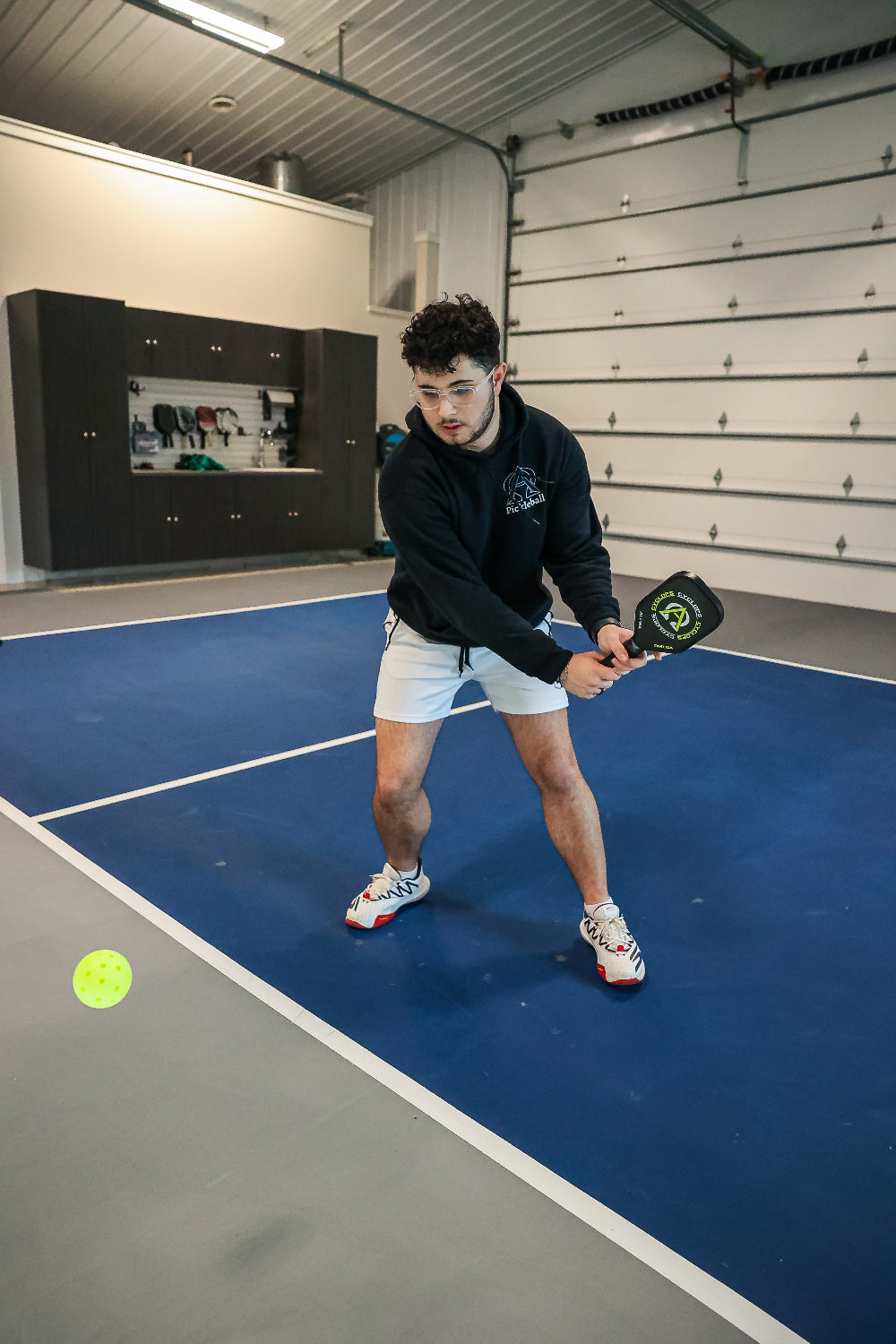 Man in black hoodie and white shorts playing pickleball indoors.