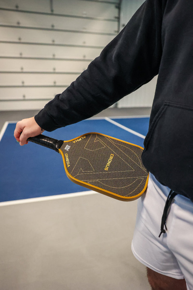 A man wearing a black hooded sweatshirt and white shorts is holding a Cronus Omega Alpha pickleball paddle.