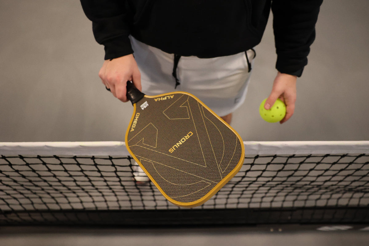 Person holding a pickleball paddle and ball over a net. They are wearing a black hoodie and white shorts.