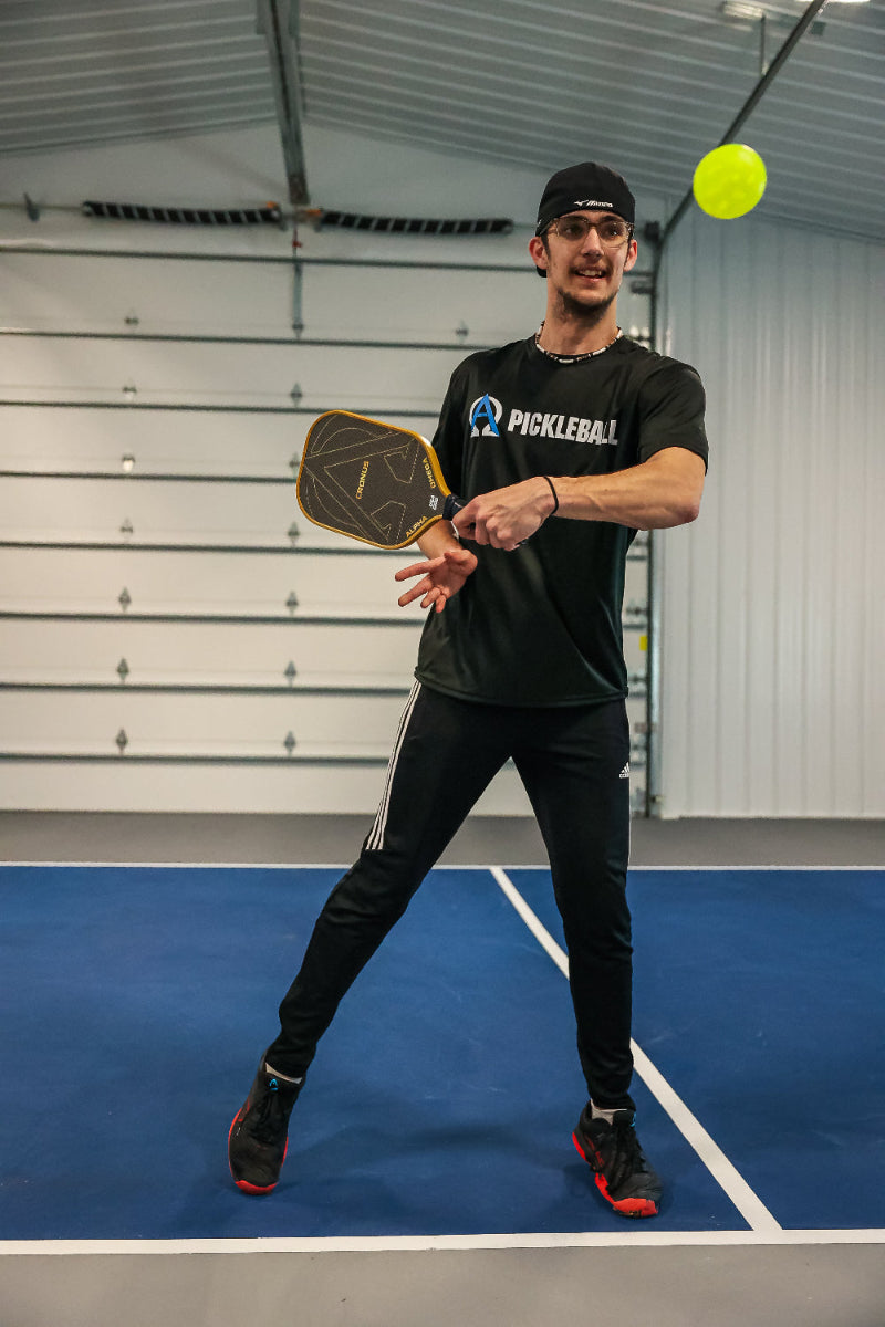 A man in a black athletic outfit playing pickleball on a blue court with a yellow ball in the air.