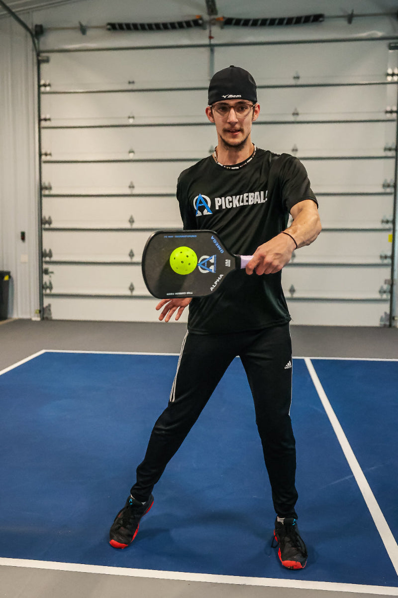 Man in black clothing holding a pickleball paddle and ball on a blue court.