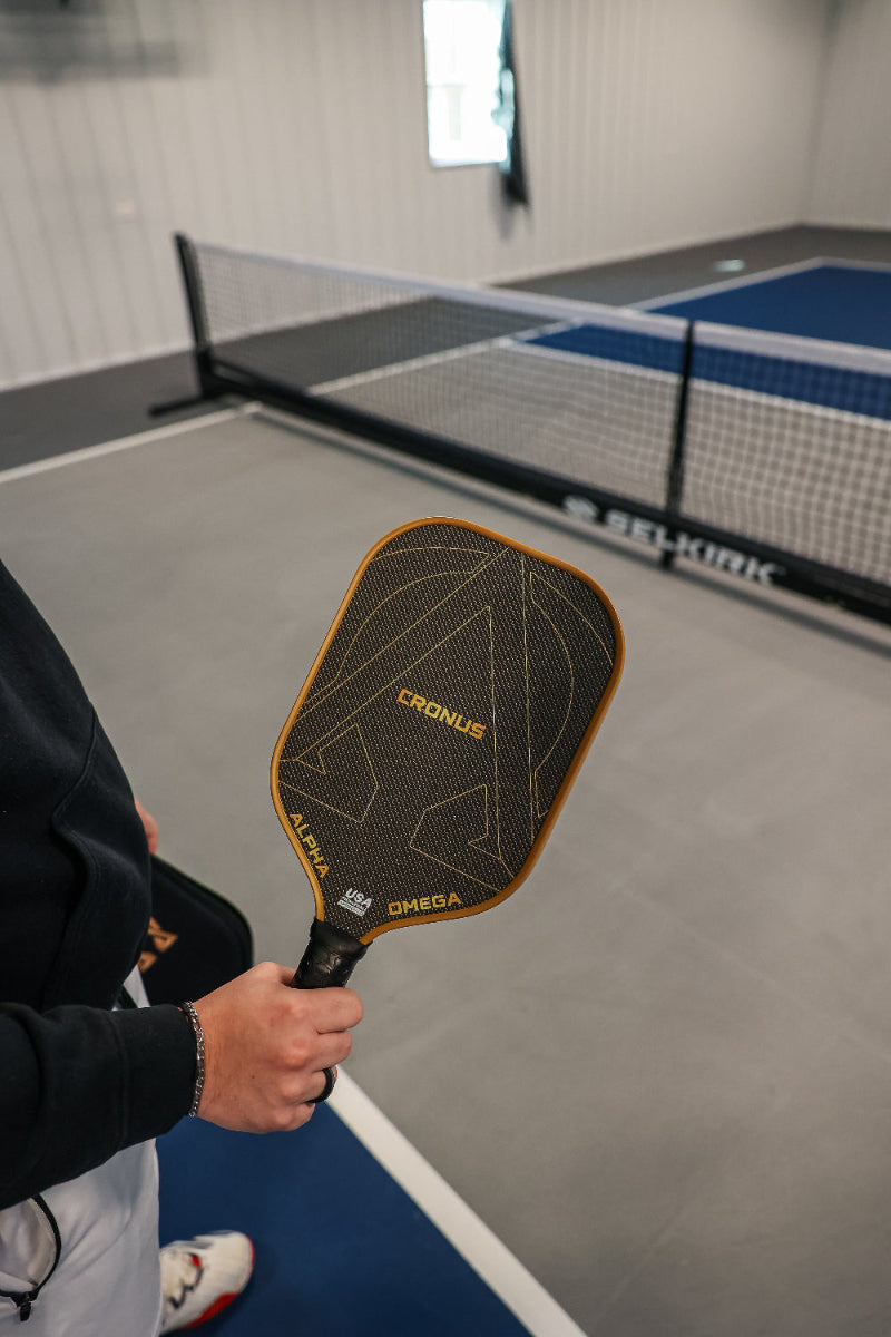 Person holding a black and gold Cronus Alpha Omega Selkirk pickleball paddle in a pickleball court setting.
