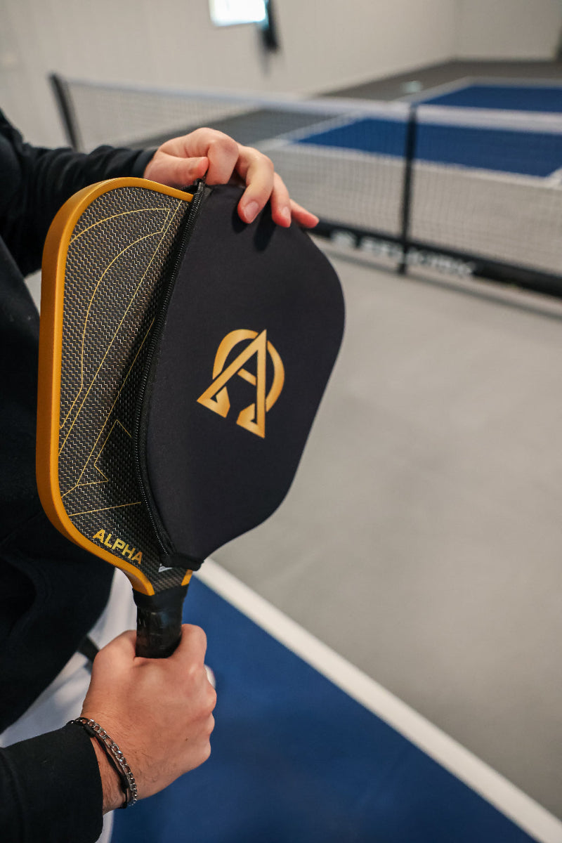 Person holding a pickleball paddle with a black cover over half of it, showing the golden net pattern on the other half, with a pickleball court in the background.