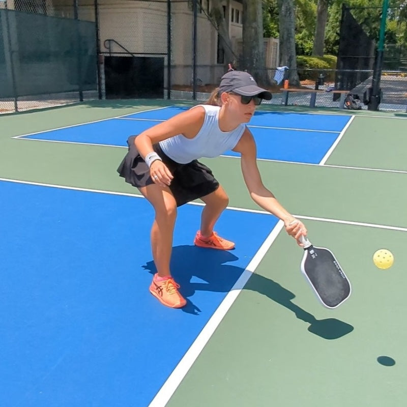 Woman playing pickleball, wearing a light blue tank top, black skirt, matching hat, sunglasses, and orange athletic shoes. She is holding a pickleball paddle, ready to hit the yellow pickleball. She is on a blue and green pickleball court.