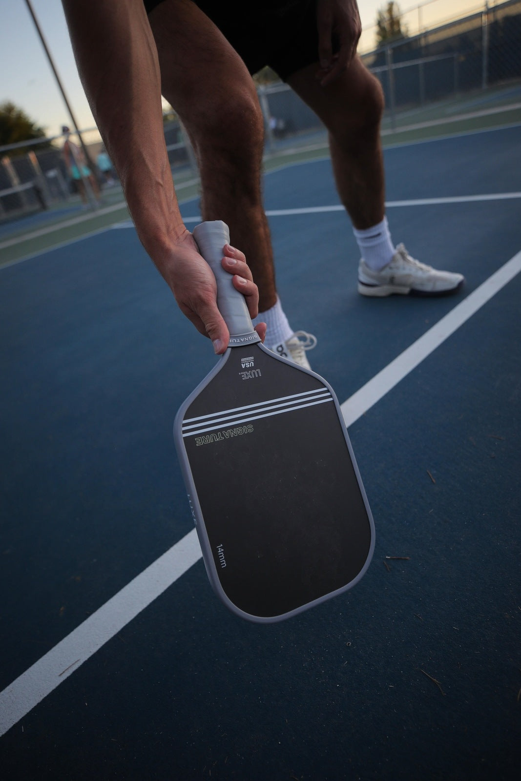Close-up of a person holding a black and gray Signature Luxe pickleball paddle on a blue court with white lines, wearing black shorts, white socks, and white sneakers.