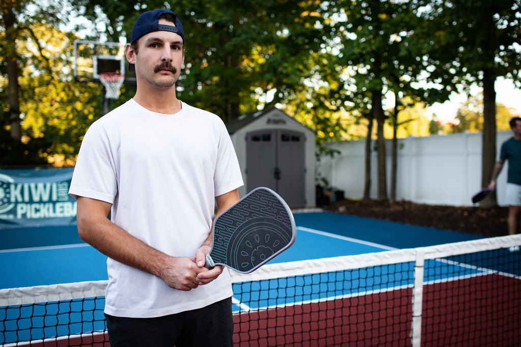 A man in a blue cap and white t-shirt holds a pickleball paddle on a blue and red court.