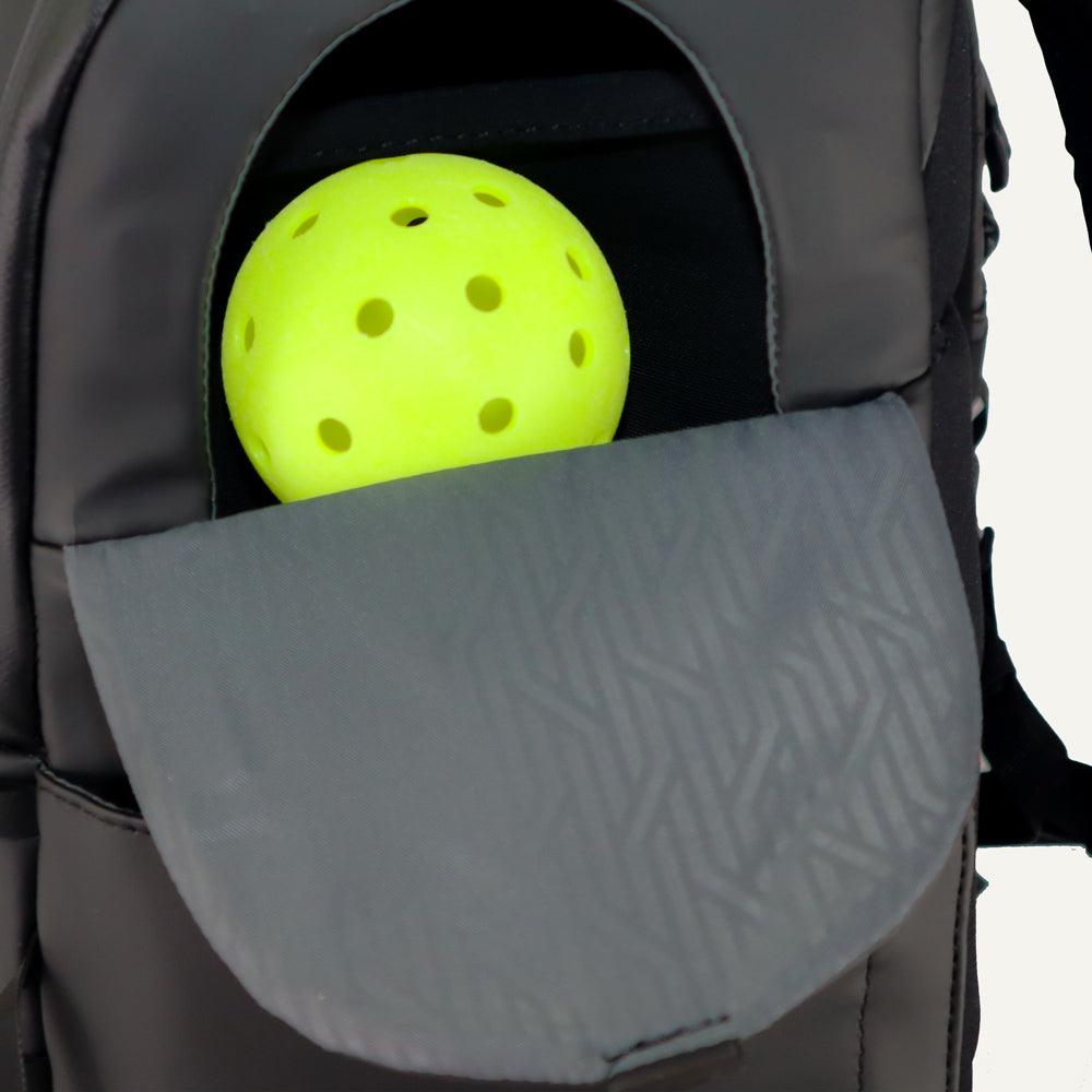 Dark gray pickleball backpack by FORWRD with an open side pocket holding a yellow pickleball and a textured flap.