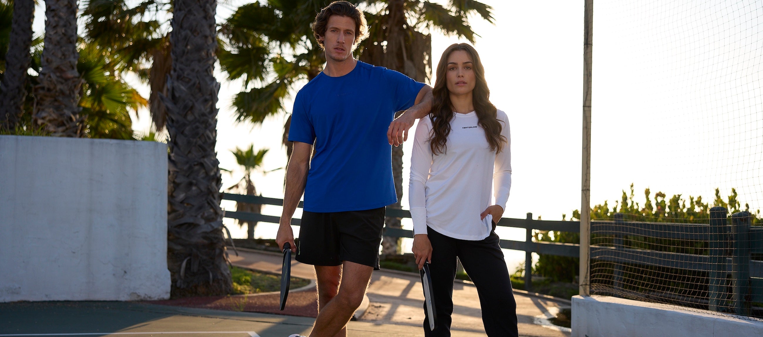 Two people wearing Centerline athletic clothing standing on a tennis court with palm trees in the background