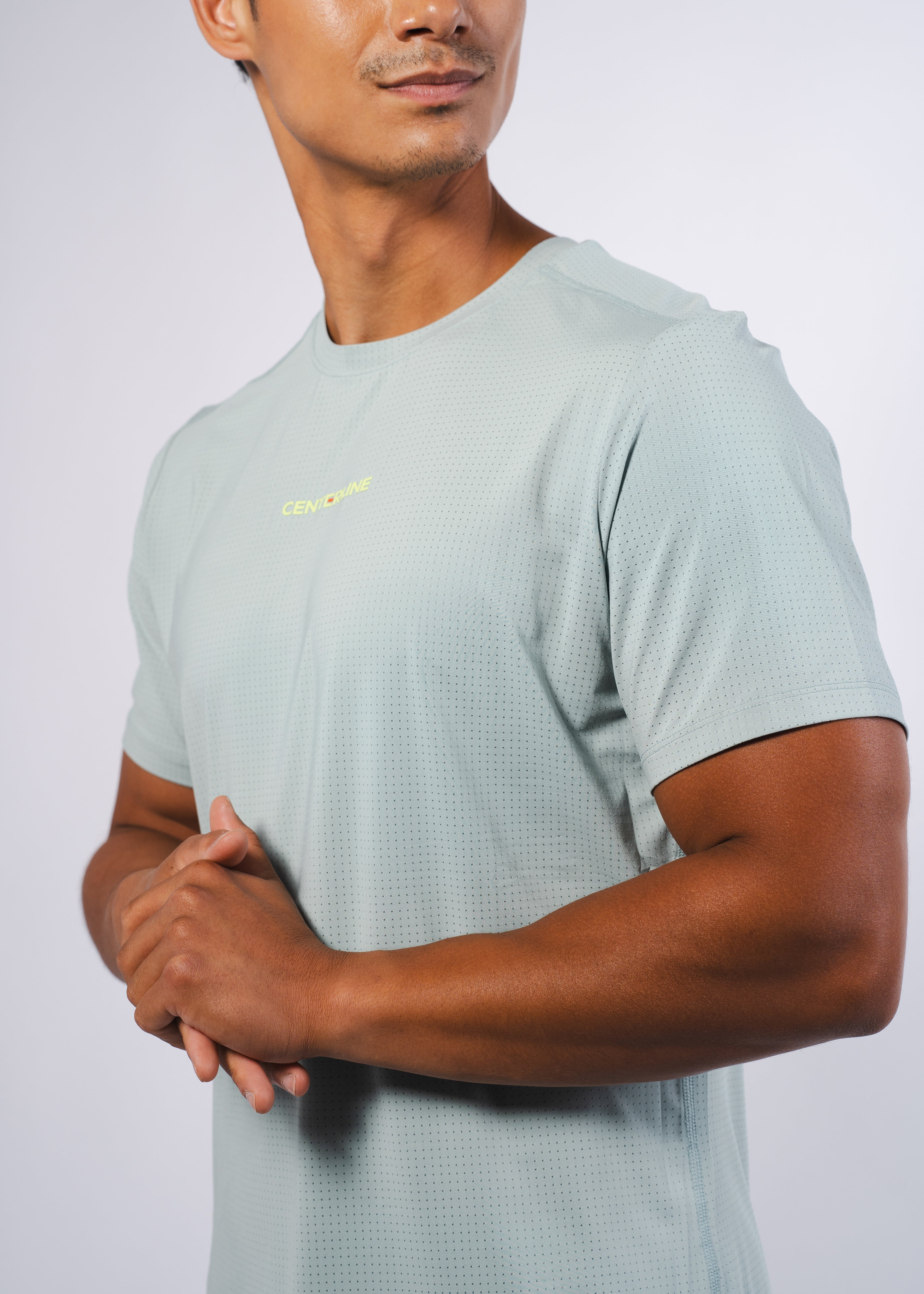 Light mint green men's perforated tee by Centerline Athletics with a yellow CENTERLINE logo, worn by a man with clasped hands.