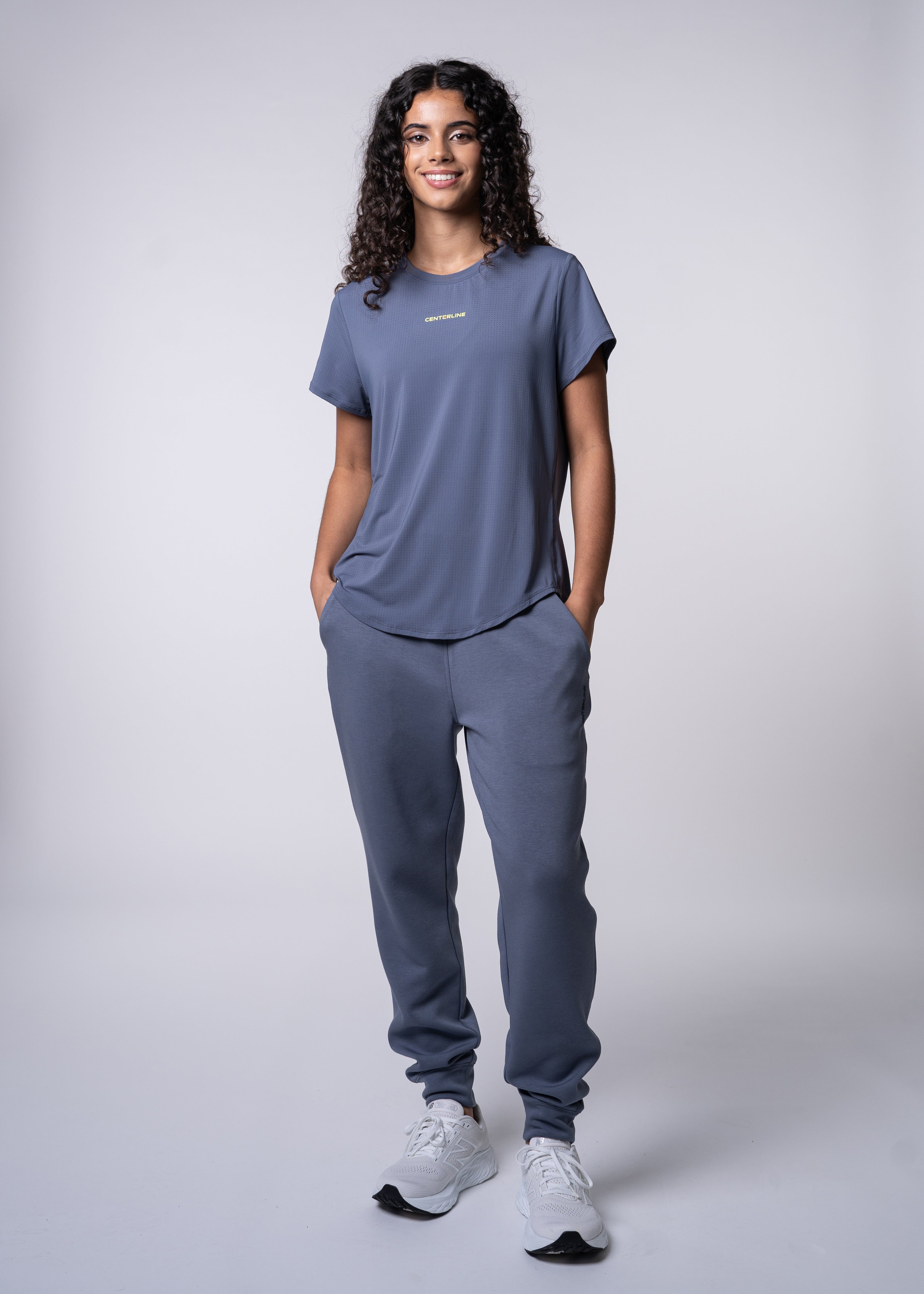 A woman in a blue t-shirt and sweatpants stands with her hands in her pockets.
