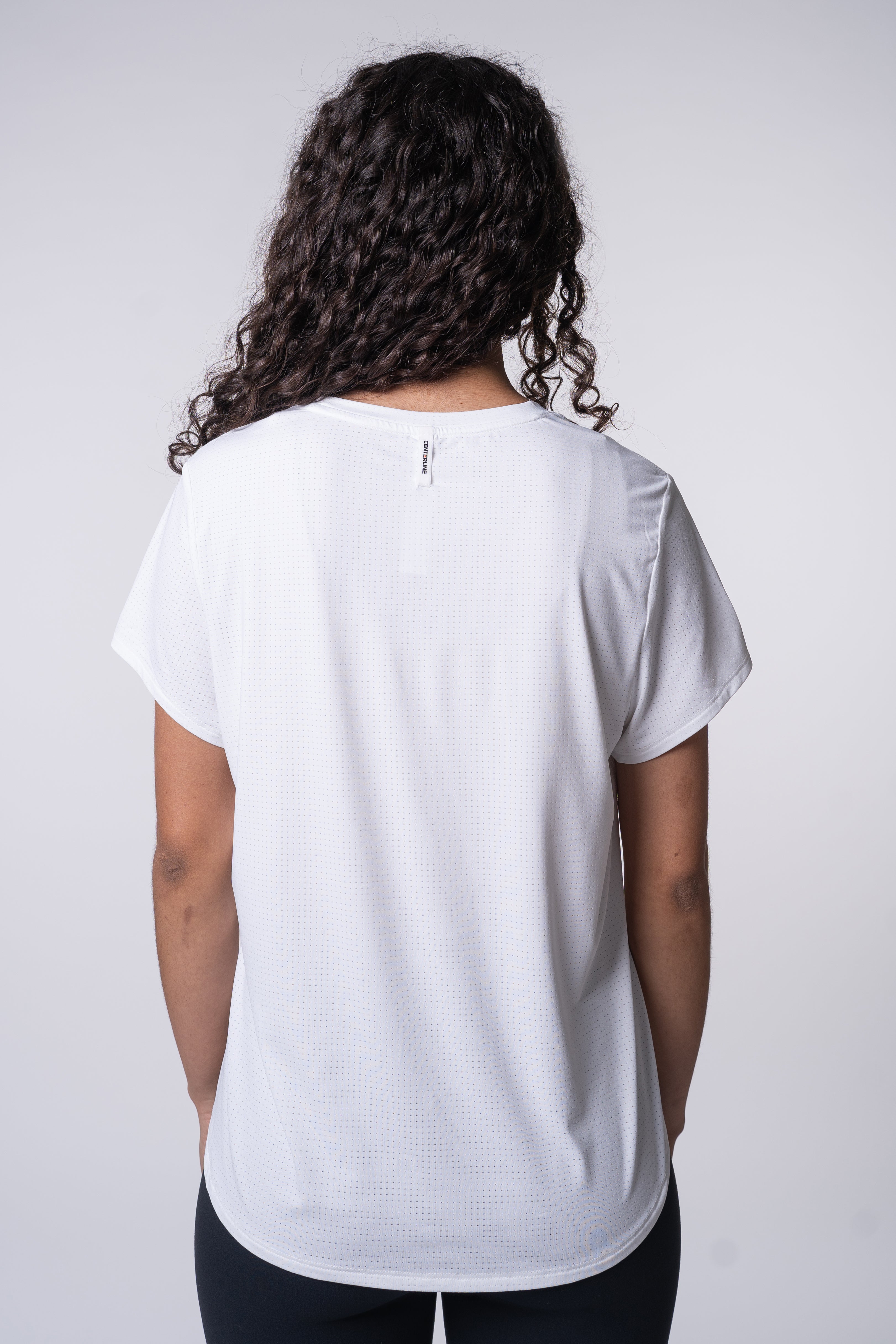 A person with curly dark hair is wearing a white athletic shirt with small holes. They are standing facing away from the camera against a white backdrop.