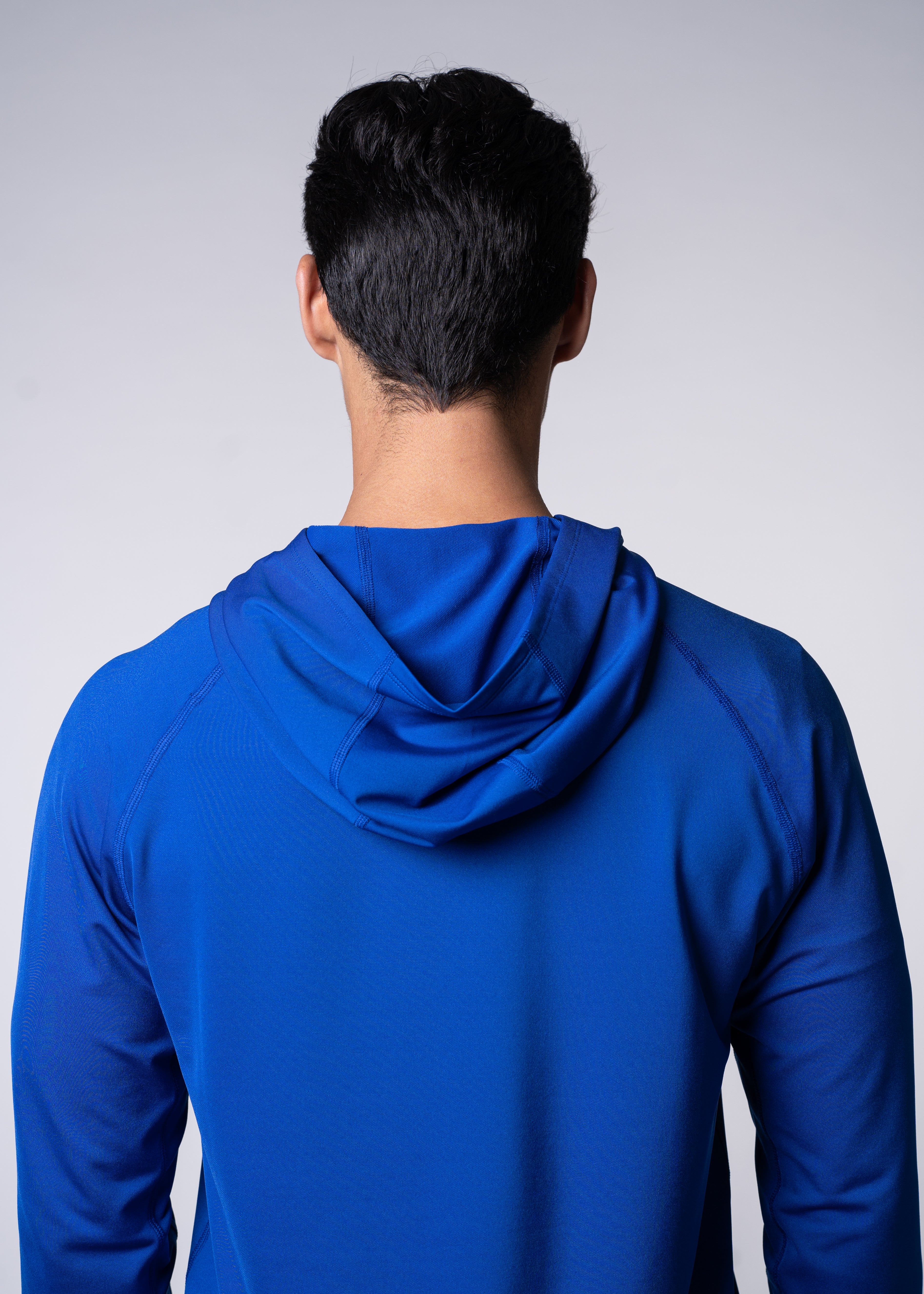 Back view of a man wearing a blue hooded long-sleeve shirt against a grey background.