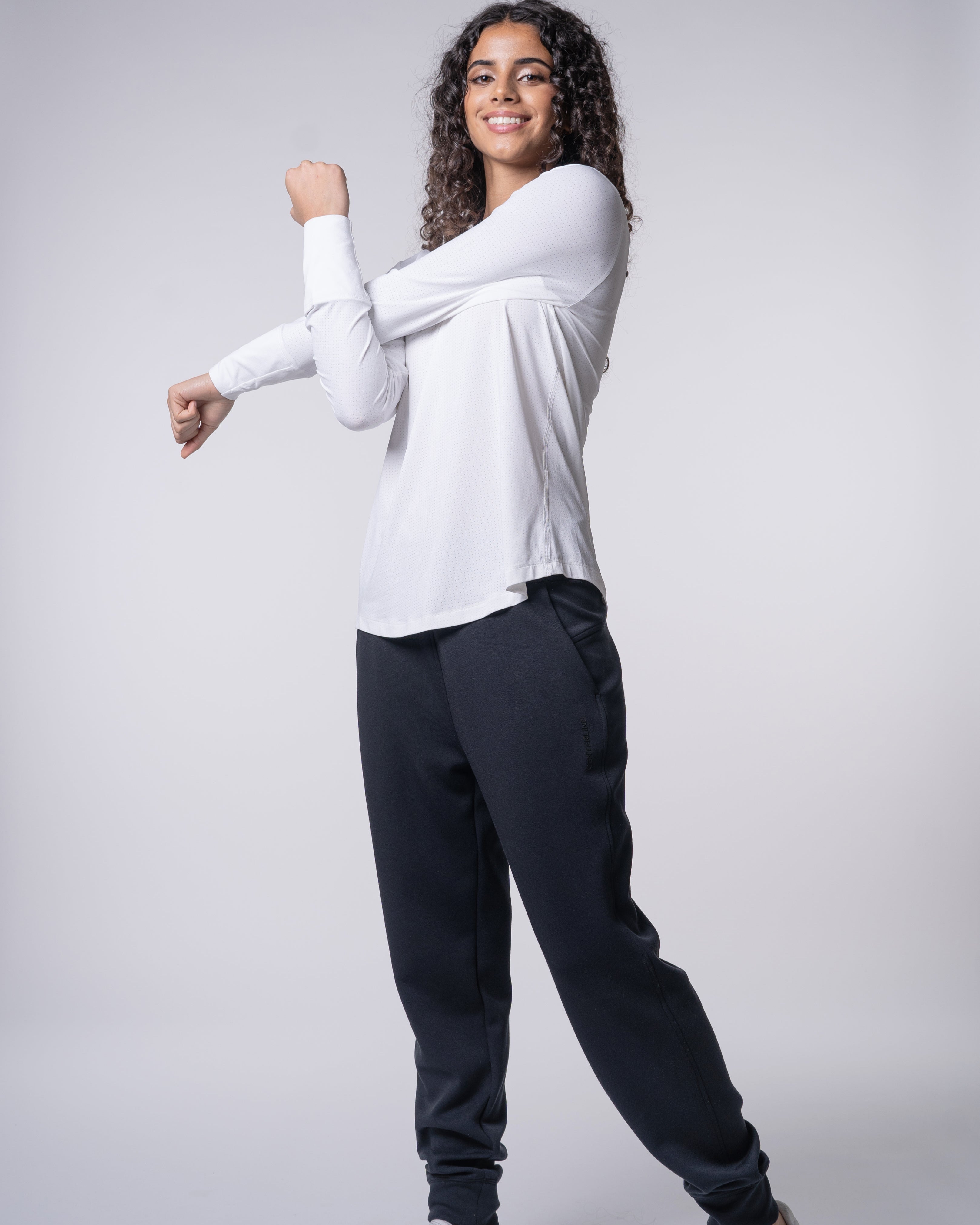 Woman in a white long-sleeve shirt and black sweatpants stretches her arms.